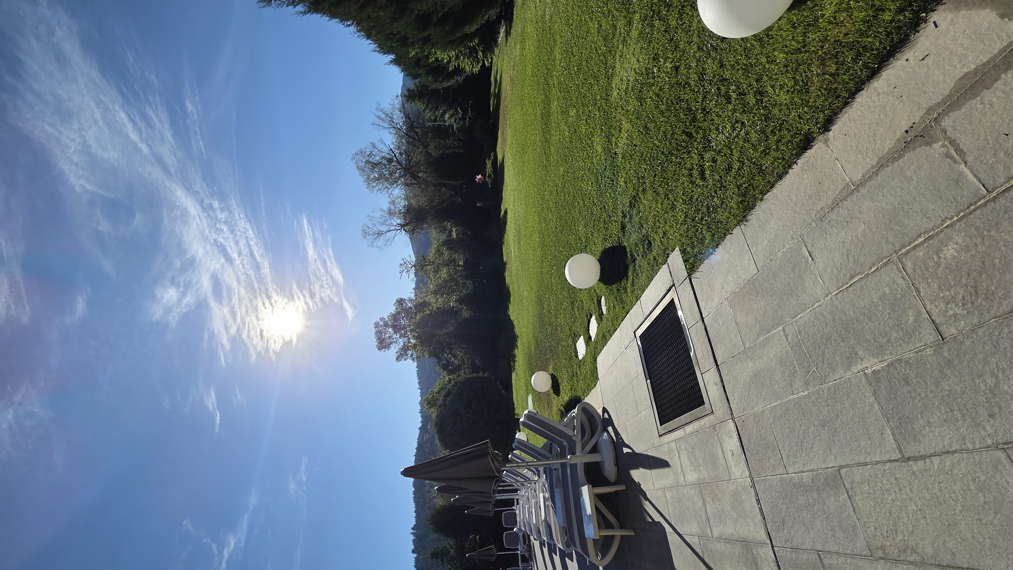 The image shows an outdoor scene with a clear blue sky and bright sun. In the foreground, there is a patio area with gray tiles. Several lounge chairs with gray cushions and dark umbrellas are neatly arranged on the patio, facing a lush green lawn. A square metal grate is embedded in the patio near the lounge chairs. The lawn is dotted with three large, white spherical lanterns. Beyond the lawn, there is a dense line of trees and hills in the background. The overall impression is of a serene and well-maintained outdoor space, likely a resort or hotel grounds.