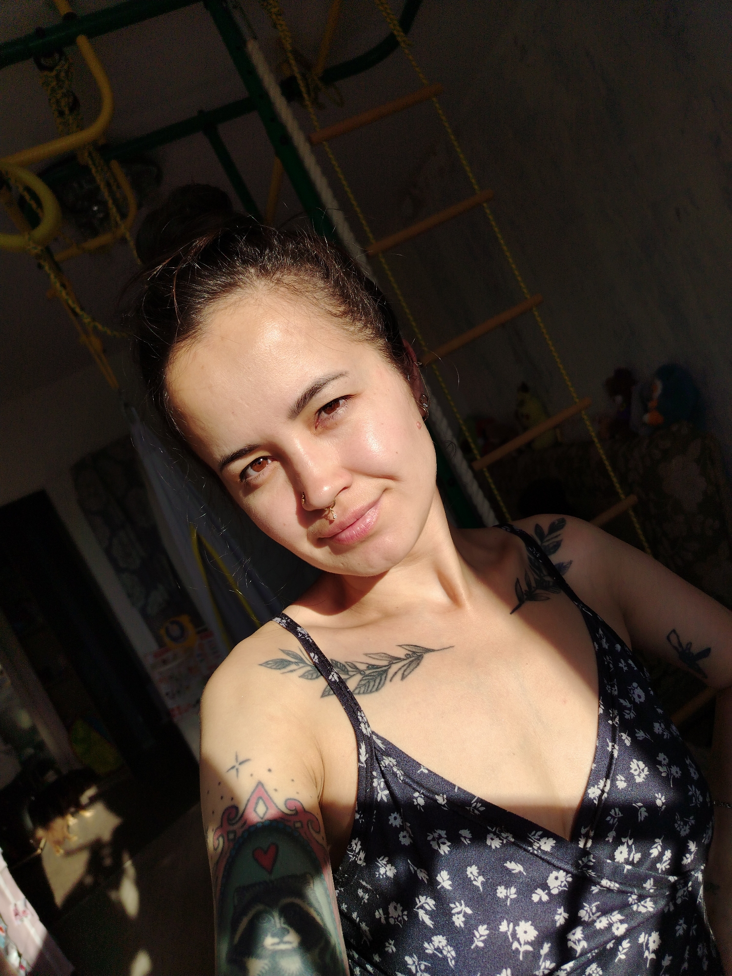 The image is a close-up, selfie-style portrait of a young woman with dark hair pulled back in a bun. She is wearing a dark blue dress with a small white floral print. She has a nose ring and several tattoos visible on her chest and arm. The lighting suggests she is indoors, possibly near a window, as sunlight is casting strong shadows. In the background, out of focus, a rope ladder and a jungle gym structure are visible, suggesting a children's play area or gym. The overall impression is informal and casual. The location within Noviy, Russia, is not discernible from the image itself.