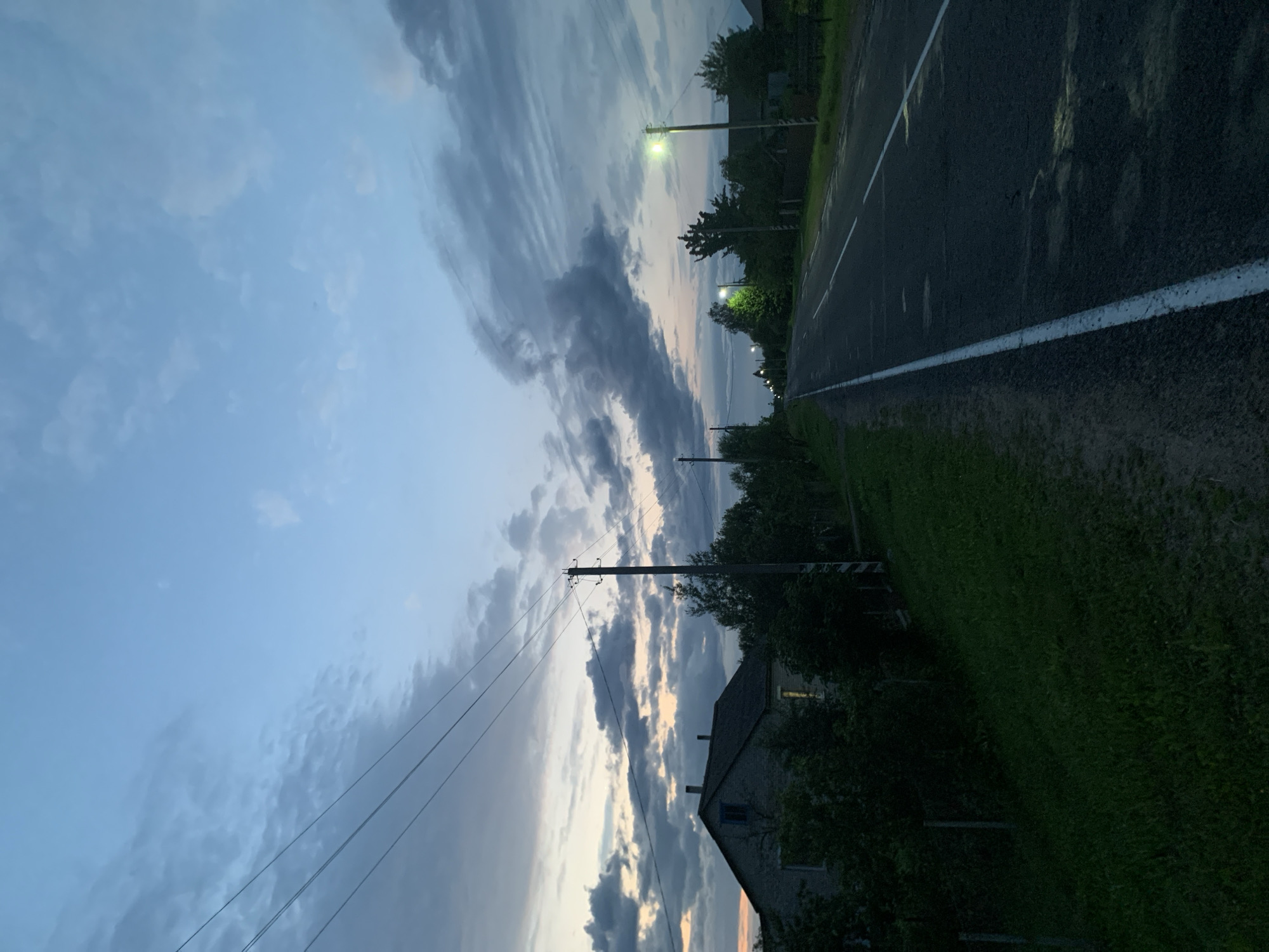 The image depicts a dusky rural scene in Відзаўскі сельскі Савет, Belarus. A paved road with a white dividing line extends into the distance, flanked by a grassy shoulder on the left and trees and houses on the right. Several streetlights illuminate the roadside. A two-story building with a dark roof and visible windows is situated to the left of the road, surrounded by dense foliage. Overhead, power lines stretch across the sky, which is dominated by a mix of light blue and dark, cloudy formations, indicating the transition from day to night. The overall atmosphere is quiet and serene.
