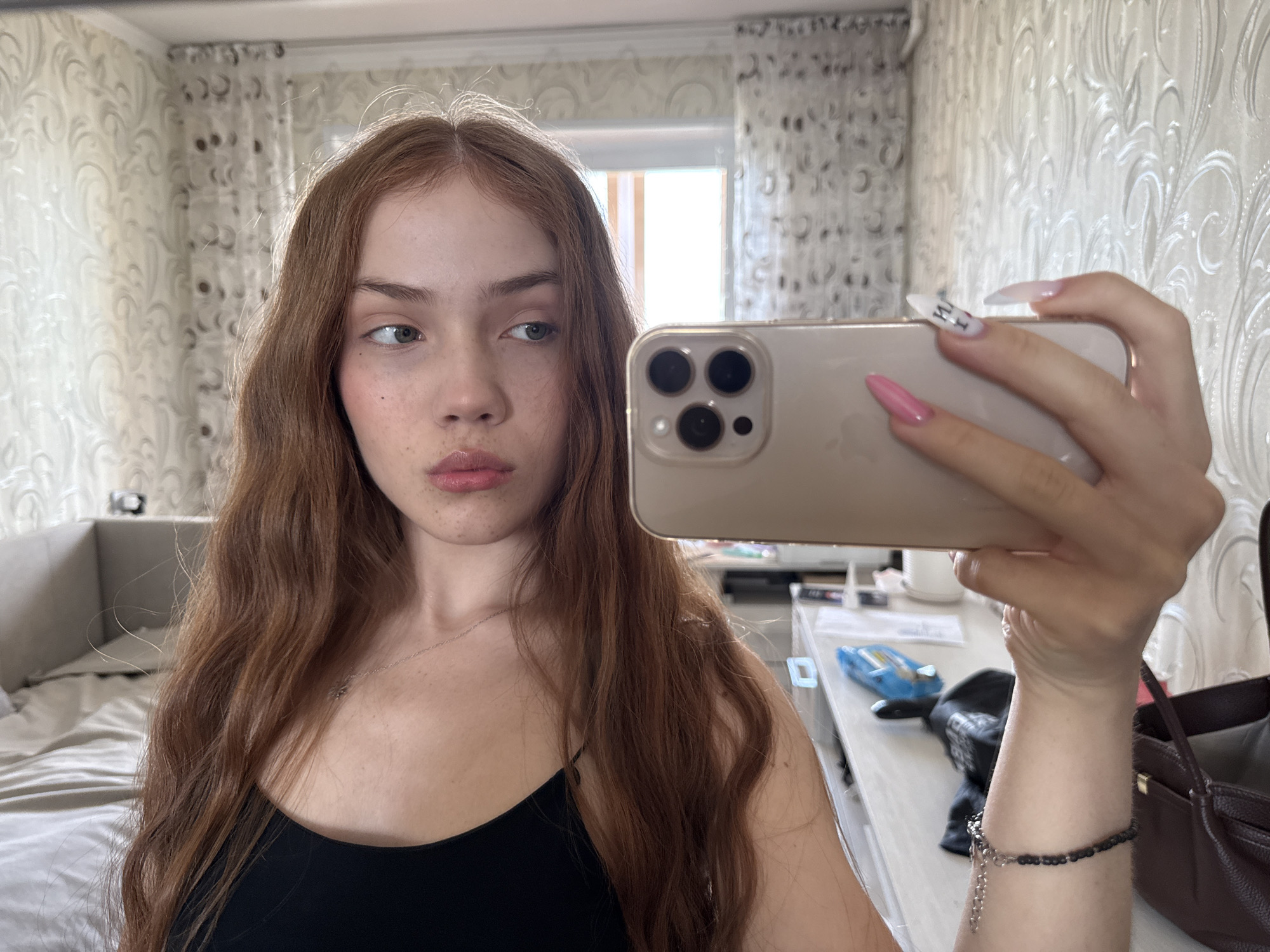A young woman with long, wavy, reddish-brown hair is captured in a mirror selfie, taken indoors in what appears to be a bedroom or living space in Naberezhnye Chelny, Russia. She wears a black spaghetti strap top and has a thin silver necklace and a dark beaded bracelet on her right wrist. Her light-colored eyes (possibly blue or green) are cast to her right, and her lips are slightly pouted. Visible freckles dot her face, particularly on her nose and cheeks.
She holds a gold-colored iPhone (likely a Pro model due to the three camera lenses) with her right hand to take the photo. Her long, pointed nails are prominent, with most painted pink and one white nail featuring partially visible black text that reads "I'M H", likely part of a phrase like "I'M HOT".
The background features light-colored wallpaper with an ornate, flowing pattern. To the left, a bed with light bedding and a headboard is visible, suggesting a bedroom. To the right, a table or desk holds various items, including a blue object resembling a toy or tool, and a dark brown handbag. Bright natural light streams in from a window, which is reflected in the background, indicating it is daytime. The overall impression is a casual, posed moment with a contemplative or slightly distant expression.