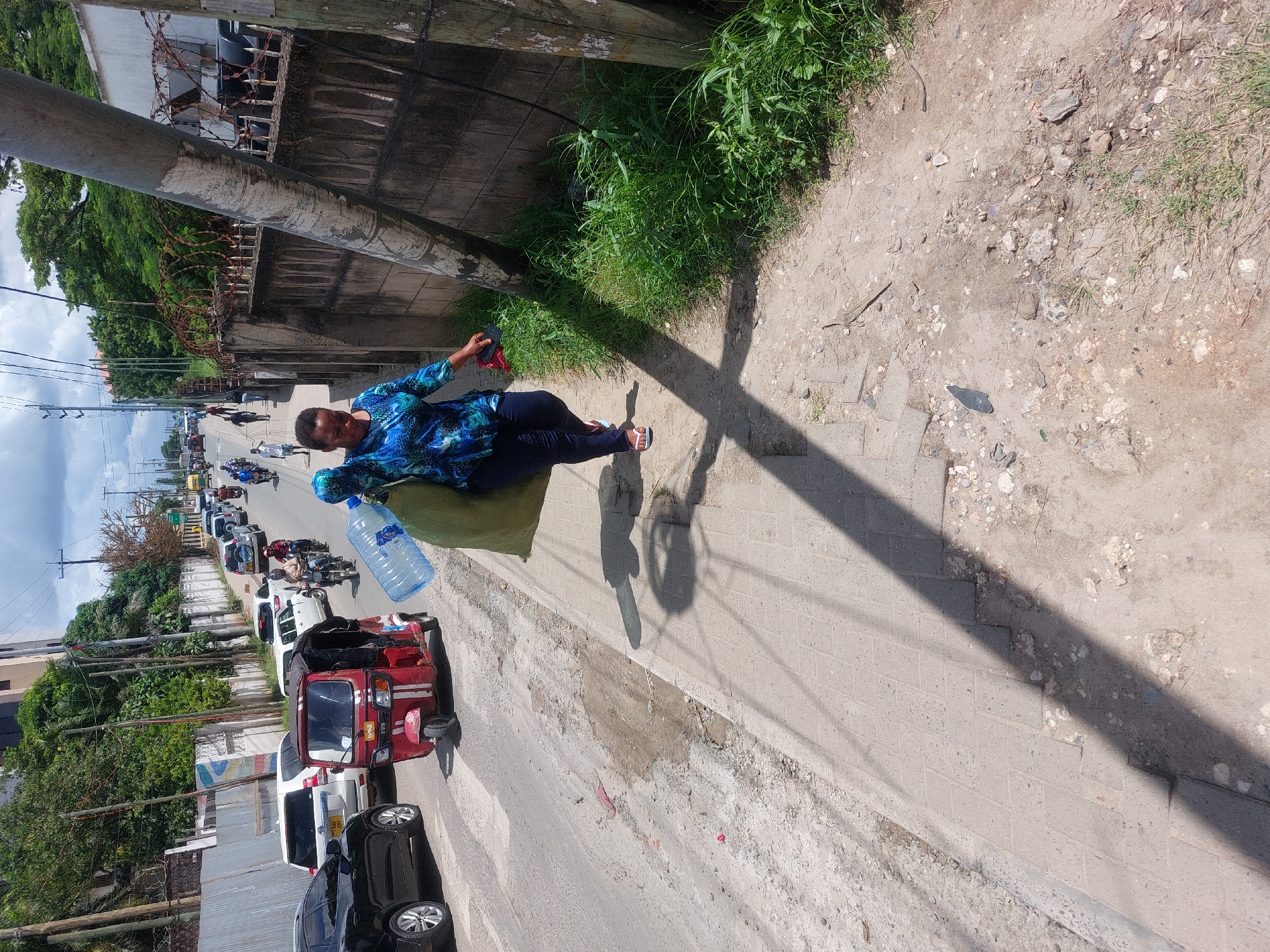 The image depicts a woman sitting on a green cushion along a sidewalk next to a road in Dar es-Salaam, Tanzania. She is wearing a blue patterned shirt and dark pants, holding a large water bottle. A red tuk-tuk is parked on the road, along with other cars and motorbikes. The sidewalk is made of concrete and has grass growing alongside it. There is a utility pole casting a shadow across the sidewalk. A tall wall with barbed wire runs along the side of the road.