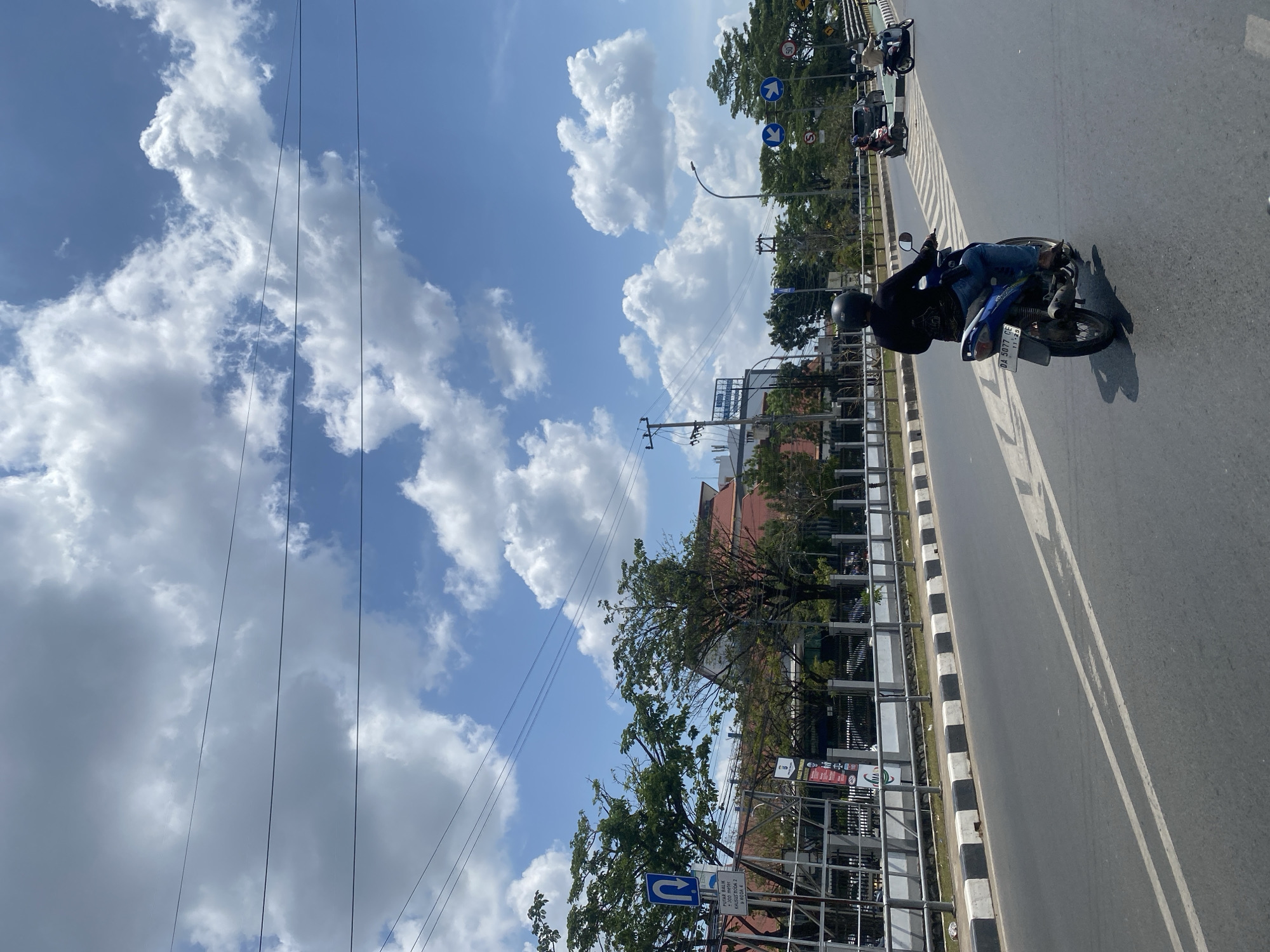 The image captures a bright, sunny day in Banjarmasin, Indonesia, with a clear blue sky dotted with fluffy white clouds. The perspective is from a moving vehicle on a road. In the foreground, a person wearing a black shirt, blue jeans, and a black helmet rides a blue motorcycle. The motorcycle has a license plate that reads "DA 5077 OE". The road is asphalt with white lane markings.
To the left of the road, there is a sidewalk bordered by a black and white striped curb. Beyond the sidewalk, a metal railing runs along a raised area. Further left, there are trees and a sign with a U-turn arrow, indicating traffic direction. In the background, a building with a reddish-orange roof is visible, along with more trees and a utility pole with wires.
On the right side of the road, other vehicles, including motorcycles and cars, can be seen. Traffic signs, including directional arrows and a speed limit sign (50), are also present. The overall scene depicts a typical urban street in Indonesia on a clear day.