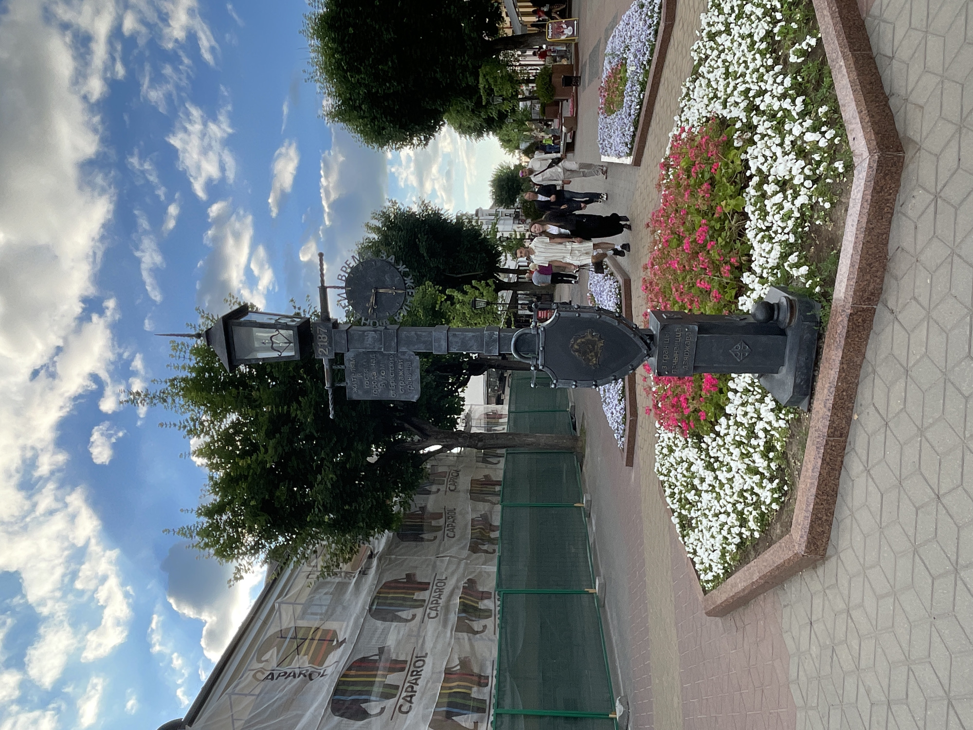 The image captures a detailed street scene on Sovetskaya Street in Brest, Belarus, under a bright blue sky with scattered white clouds. A prominent dark grey decorative lamppost stands in the central foreground, featuring a glass-enclosed lantern at its apex, a circular black clock face with Roman numerals and no visible hands, and several signs displaying Cyrillic text, including the number "2187" and a shield-shaped emblem.
The base of the lamppost is integrated into an octagonal flowerbed, bordered by a reddish-brown granite curb. This flowerbed contains a dense arrangement of white and pink flowering plants. Further down the promenade, another flowerbed with purple flowering plants is visible. The ground is paved with interlocking pavers forming geometric patterns in shades of grey and reddish-brown.
To the left of the lamppost, a multi-story building is partially obscured by white scaffolding netting. This netting is adorned with the brand name "CAPAROL" and repeated stylized illustrations of elephants in various striped color patterns. A green construction fence runs horizontally in front of this building.
To the right and extending into the background is a pedestrian promenade lined with mature green trees. Several individuals are present on the promenade. In the mid-ground, a group of four people walks towards the viewer: a man in a light striped shirt and shorts, a woman in a black long-sleeved top and skirt, an individual in a dark jacket, and a man in light trousers and a light shirt wearing a baseball cap. Other pedestrians are visible further in the distance. Buildings with commercial establishments are present along the promenade, with a partially visible sign displaying "ПАРФЮМЕР" (Perfumer) on the right side of the street.
