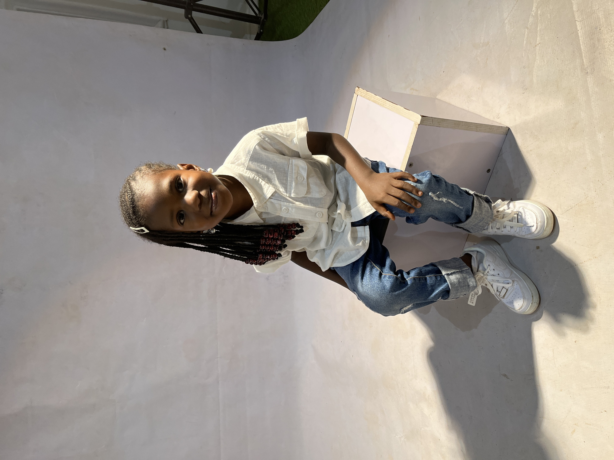 A young girl with dark skin, wearing a white button-down shirt, blue jeans, and white sneakers, sits on a white box indoors against a white backdrop. She has dark braided hair with beads and a silver barrette. The location is likely a photography studio or a similar setting in Jos, Nigeria, given the mention of the city. The environment appears to be artificially lit.