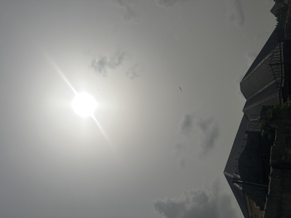 This outdoor image presents a bright, hazy sky over the city of Obinze, Nigeria. A prominent, intensely luminous sun with a distinct starburst effect occupies the upper-center to upper-left quadrant, dominating the sky. Scattered patches of light to dark grey clouds are visible, primarily in the lower-left and upper-right areas. Several dark avian figures are observed in flight against the sky, including one larger bird with outstretched wings in the middle-right. The lower third of the frame shows the dark silhouettes of multiple building rooftops, characterized by geometric, likely pyramidal or hip designs, constructed with dark-colored materials, possibly corrugated metal. A substantial dark-colored concrete or stone wall runs across the bottom of the image, with decorative metal railings visible atop sections of it and between rooflines. Sparse, dark green foliage is visible growing near the wall. No human figures or direct human activities are present.