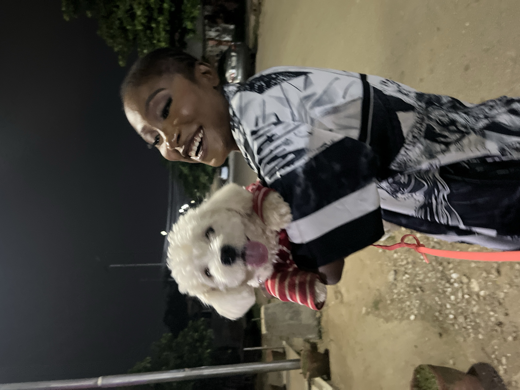 A black woman with short hair and bold makeup is holding a small white dog. She is laughing and looking up and to her left. She is wearing a black and white patterned jacket. The dog is wearing a red and white striped shirt. A red leash is visible. They are outdoors at night. The background shows a street, trees, and a parked car. The scene appears to be in a residential area. The woman's excited expression and the dog's playful demeanor suggest a positive interaction. The lighting indicates nighttime, possibly from streetlights or artificial illumination. The environment suggests an urban or suburban setting within Abuja, Nigeria.