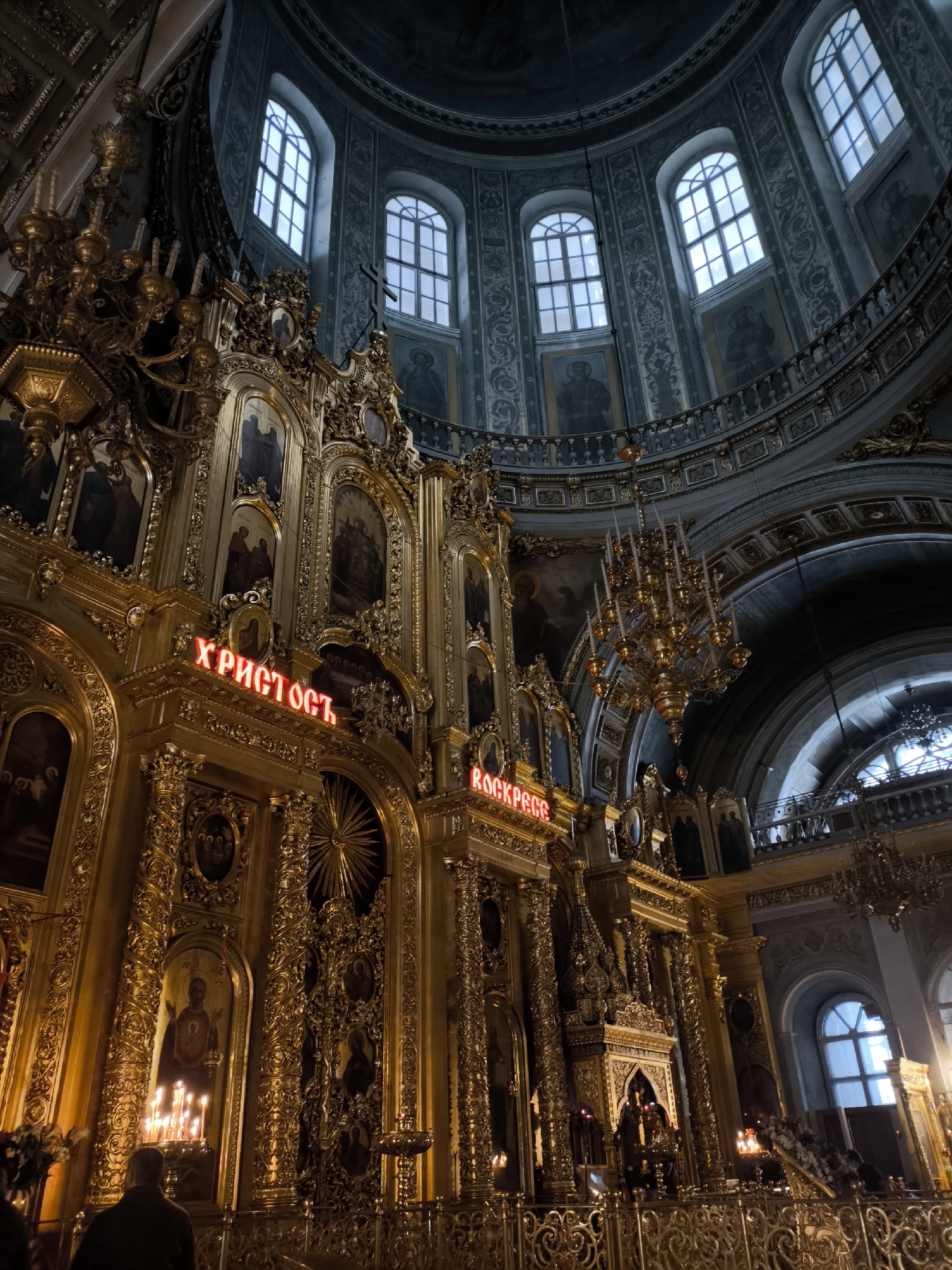 The image depicts the interior of a cathedral in an unidentified city. The architecture features a large, ornate golden iconostasis with lit candles and illuminated Cyrillic script reading "ХРИСТОСЪ" and "ВОСКРЕСЕ." Above the iconostasis, the cathedral's dome rises, with a series of arched windows and decorative paintings. Chandeliers hang from the ceiling. The overall environment suggests a grand, religious setting with a blend of natural and artificial light. An individual is partially visible in the foreground.
