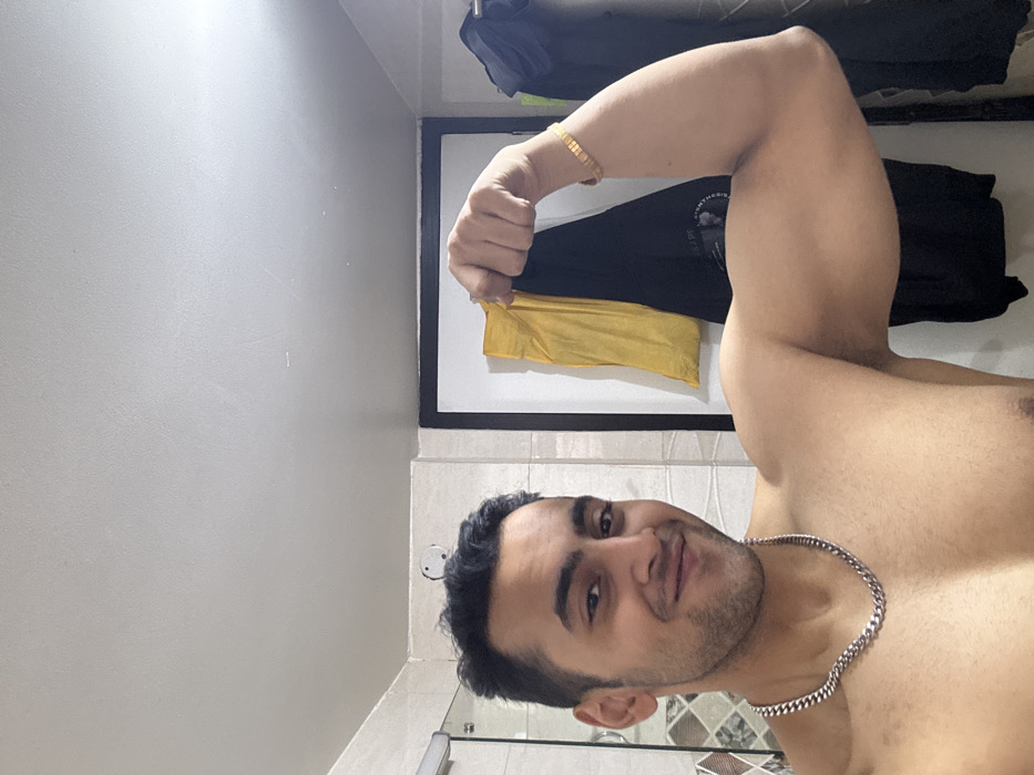 A shirtless man flexes his right bicep, displaying its size. He wears a gold bracelet on his right wrist and a silver chain around his neck. He faces the camera with a slight smile. Behind him, hung on a rack, are dark-colored clothes and a yellow garment. The background suggests a bathroom setting, with a tiled wall and a mirror visible below the chest.