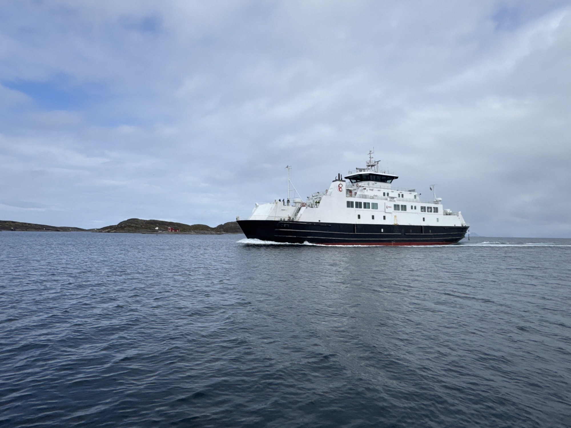 A black and white car ferry with a red stripe along its waterline sails through choppy blue water under a cloudy sky. The ferry, with its visible decks and bridge, is in motion, creating a wake behind it. In the distance, a small, rocky island with a few red buildings and a white structure is visible. The overall scene depicts a nautical journey in a coastal environment.