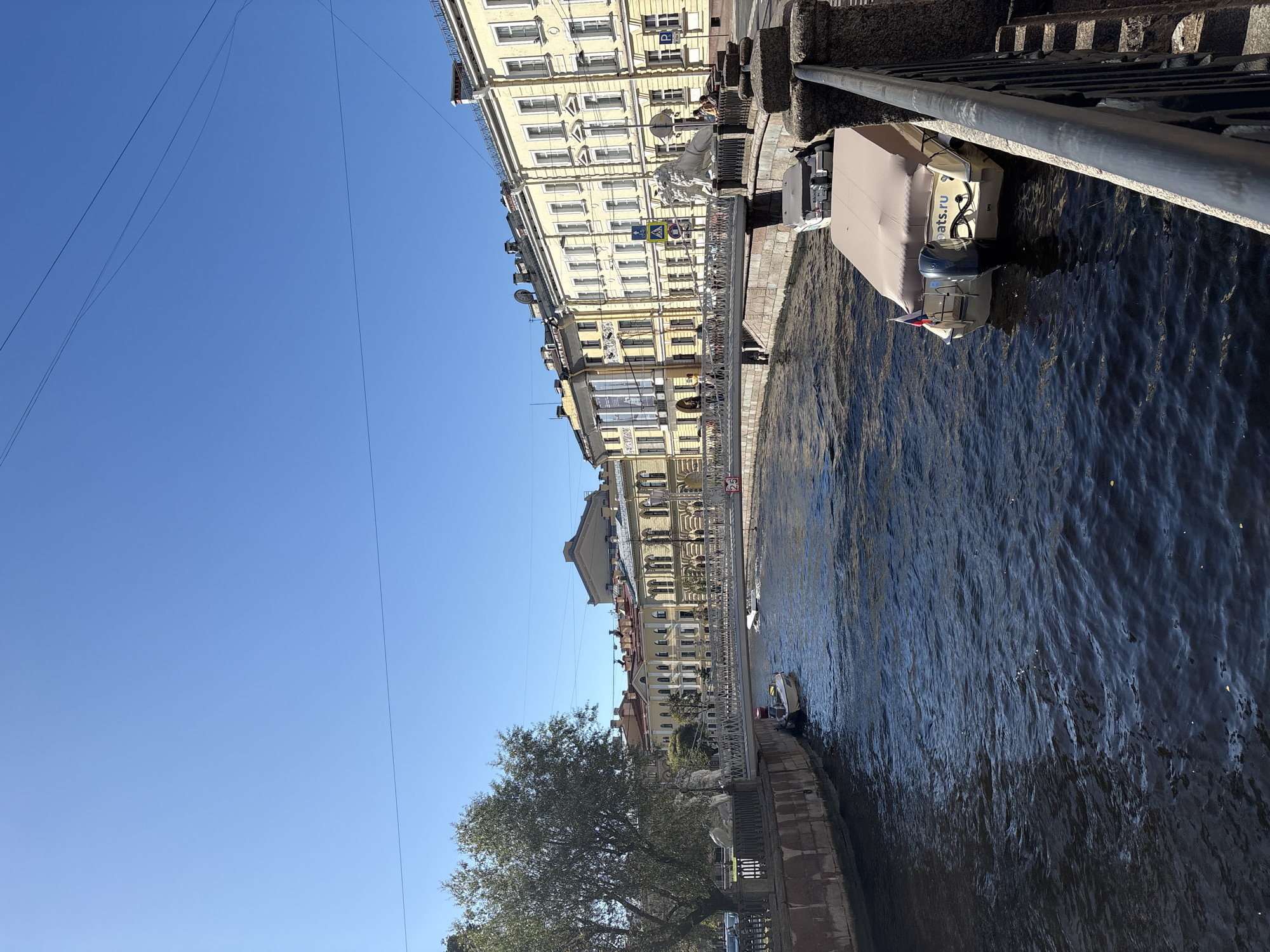 The image captures a view of the Griboyedov Canal in Saint Petersburg, Russia, on a clear, sunny day. In the foreground, dark, rippling water occupies the lower portion of the frame, reflecting the sky. A black metal railing runs along the right side, partially obscuring a stone embankment. Two boats are visible on the water: one in the mid-ground, covered with a tan tarp, and another smaller boat further back, also covered.
The canal is spanned by a pedestrian bridge with a white metal railing, where several figures can be seen walking. On the left bank of the canal, a large, leafy tree stands beside a stone embankment topped with a fence. A statue of a lion is visible on this embankment, near the bridge.
The background is dominated by a row of pale yellow buildings with multiple windows and classical architectural details. These buildings line the far bank of the canal. The sky above is a bright, cloudless blue. Overhead power lines are visible against the sky. The overall scene depicts a typical urban landscape along a canal in Saint Petersburg.