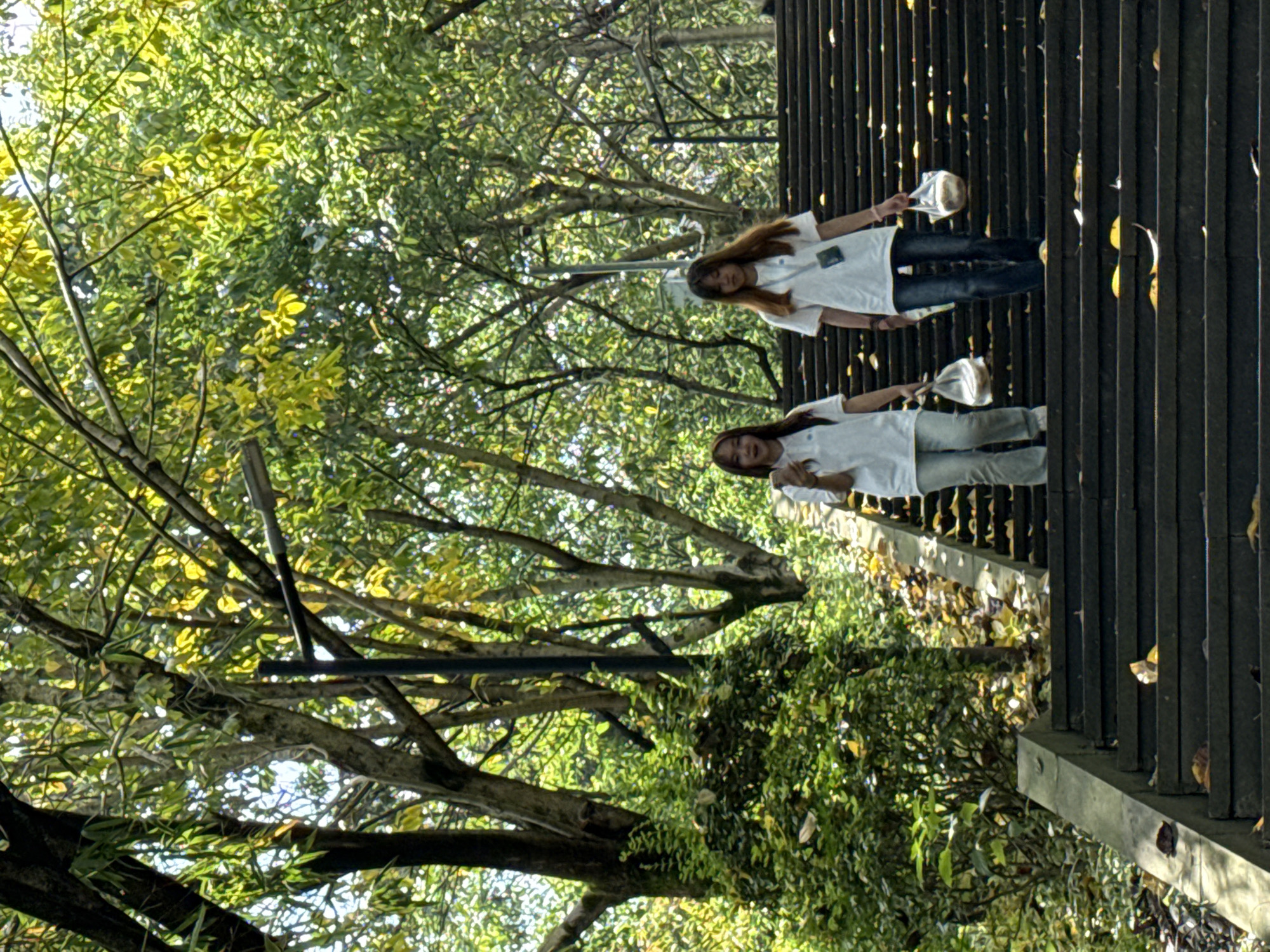 Two young women are standing on an outdoor staircase surrounded by trees in Jiangjin, China. They both wear white t-shirts, jeans, and hold small foil packets. The staircase is constructed of dark metal bars. Leaves and debris are scattered along the stairs and the adjacent ground. The surrounding environment is filled with lush trees.