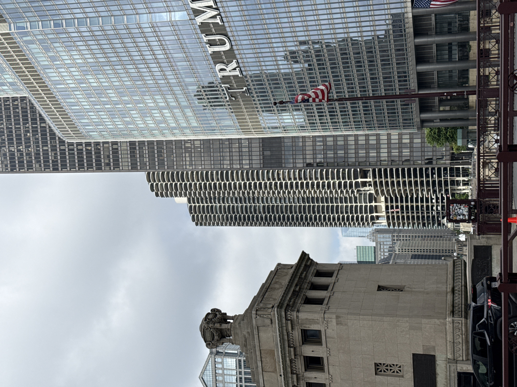 This is a vertical, outdoor photograph taken in a city. The main subjects are several tall buildings, with varying architectural styles. In the foreground, a stone building with classical detailing and ornate carvings is visible. Behind and above it, a very tall, modern building with a grid-like glass facade dominates the upper portion of the image. The words "TRUMP" are prominently displayed in large, white lettering on this glass building, along with an American flag. To the left of the Trump building, another skyscraper with a darker, gridded facade can be seen. Further back and to the left, a curved, multi-story building with a distinctive repeating pattern of balconies or windows adds to the urban landscape.
The sky is overcast, suggesting a cloudy or potentially humid day. The time of day is difficult to ascertain precisely due to the lighting conditions, but it appears to be daytime. The presence of the Trump building and the architectural styles of the surrounding skyscrapers strongly suggest the location is Chicago, Illinois. There are no people clearly visible in the frame, but hints of a street scene, including a parked car and metal railings, are present on the right side of the image. The overall impression is one of an urban environment with a mix of historic and modern architecture.