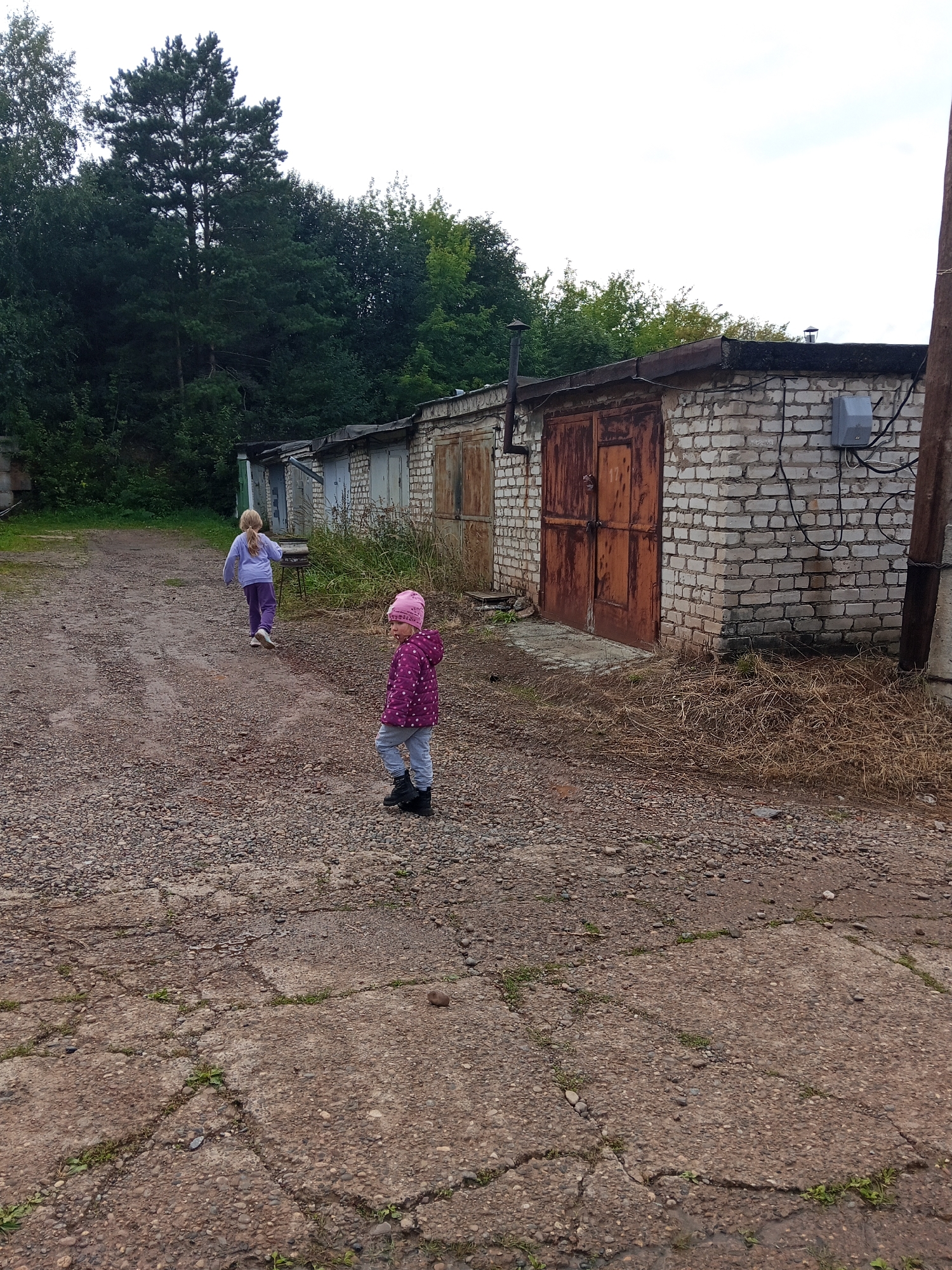 Two young girls are standing in a gravelly, cracked asphalt area next to a row of garages in the city of Chaikovsky, Russia. The girl in the foreground is wearing a pink polka-dotted jacket, a pink hat, gray pants, and black boots. She is looking to her left. The second girl, further back and to the left, is wearing a purple long-sleeved shirt and purple pants. She is walking away from the camera, towards the garages. The garages are made of white brick with metal doors. Trees and vegetation are visible behind the garages. The sky is overcast.