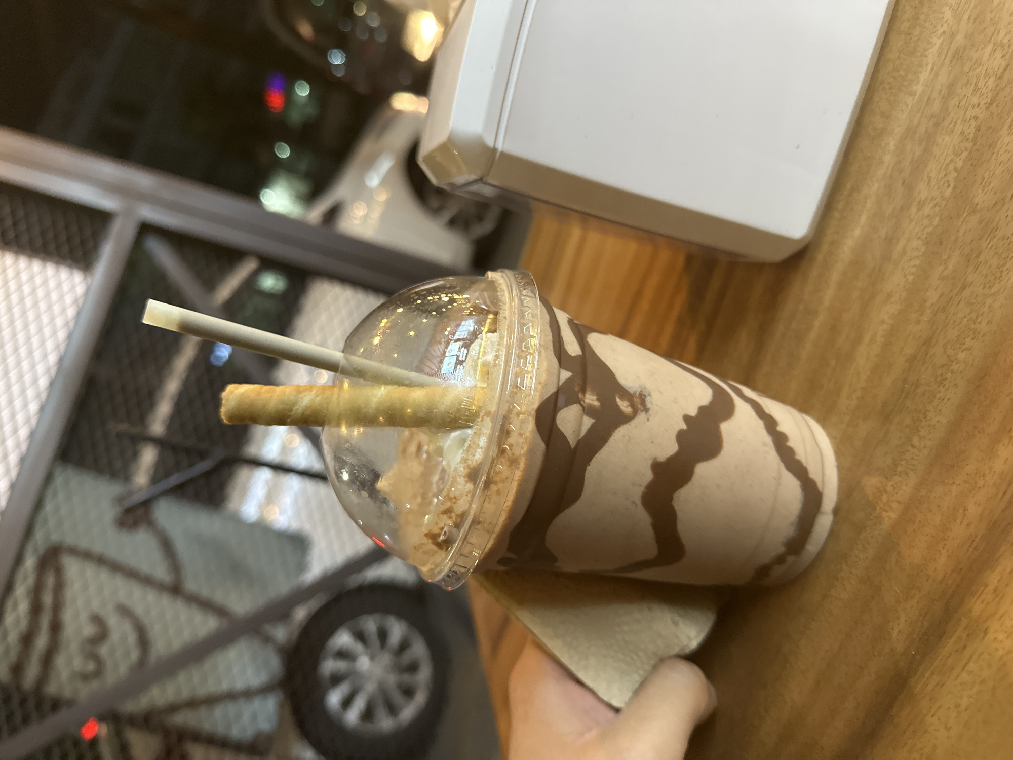 The image depicts a chocolate milkshake in a clear plastic cup with a dome lid. The milkshake is decorated with a chocolate swirl pattern. A rolled wafer stick and a straw are inserted into the milkshake. The milkshake sits on a wooden table. A person's hand is visible holding a napkin under the cup. In the background, there is a metal frame with a blurred cityscape visible through it. Based on the signage and architectural styles visible, the location is likely Puebla, Mexico.