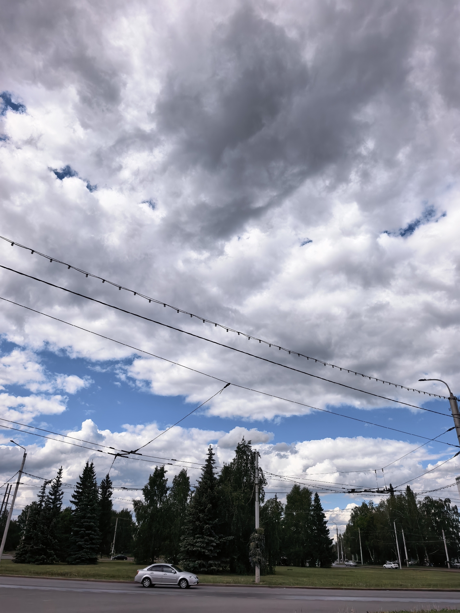 A photograph taken in Sterlitamak, Russia shows a street scene under a dramatic sky. The upper portion of the frame is dominated by layered clouds, ranging from bright white to dark gray. Below the clouds, a network of overhead power lines stretches across the scene, supported by utility poles on either side of the frame.
The lower third of the image depicts a street with a sidewalk and grassy median. Trees of varying types line the median. A silver car is parked along the edge of the road.
The lighting suggests it is daytime, with sunlight breaking through the clouds. The scene is static and devoid of visible human activity, other than implied by the presence of the car and the built environment.