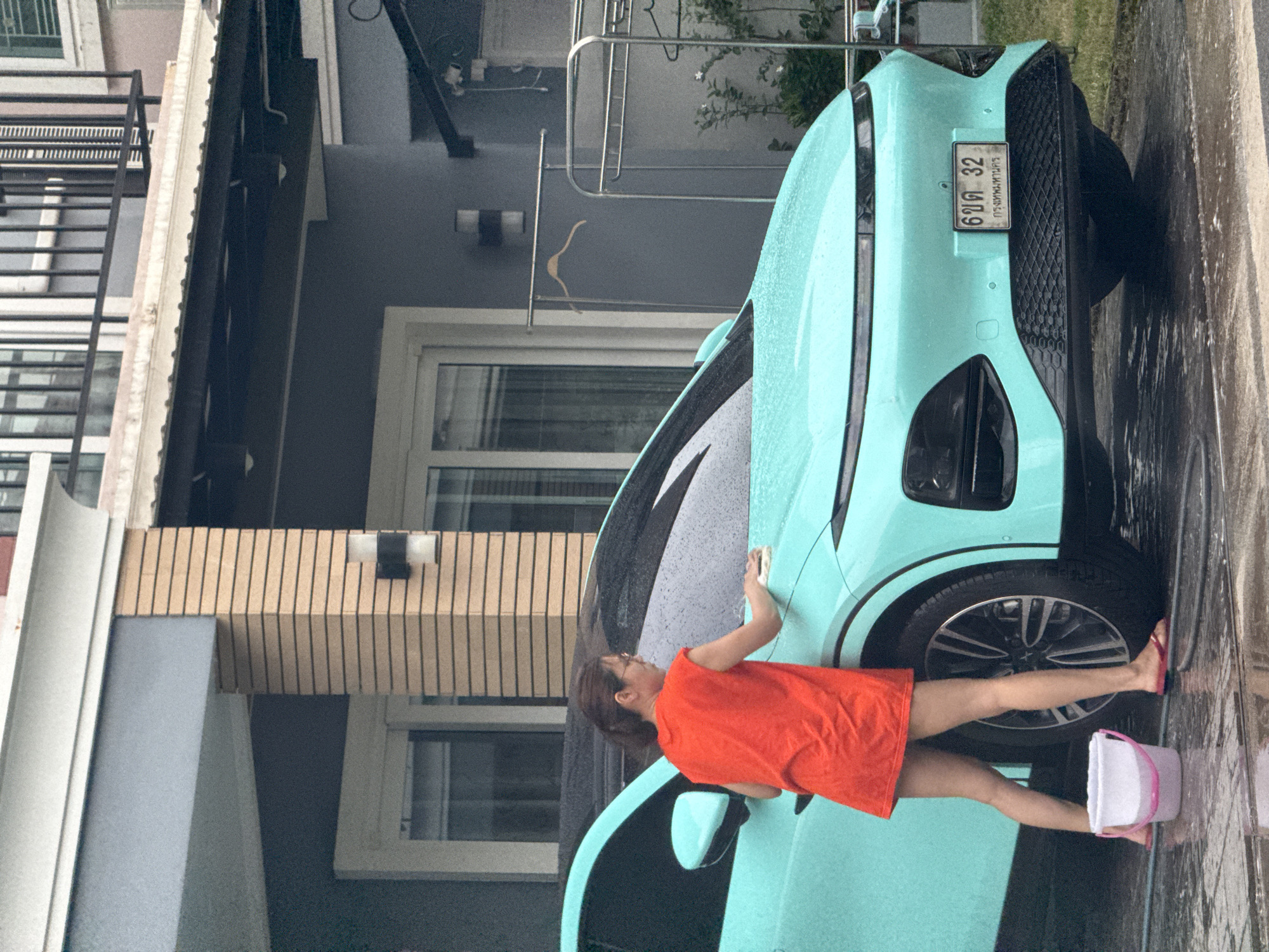 A woman in an orange dress is washing a light blue electric car in front of a house in Bangkok, Thailand. The car has a license plate that reads "6 ขด 32 กรุงเทพมหานคร". The ground is wet, indicating that it is either raining or the car is being washed. There is a bucket of water next to the car, and a clothes rack in the background. The house appears to be modern with a light-colored facade and dark gray accents.