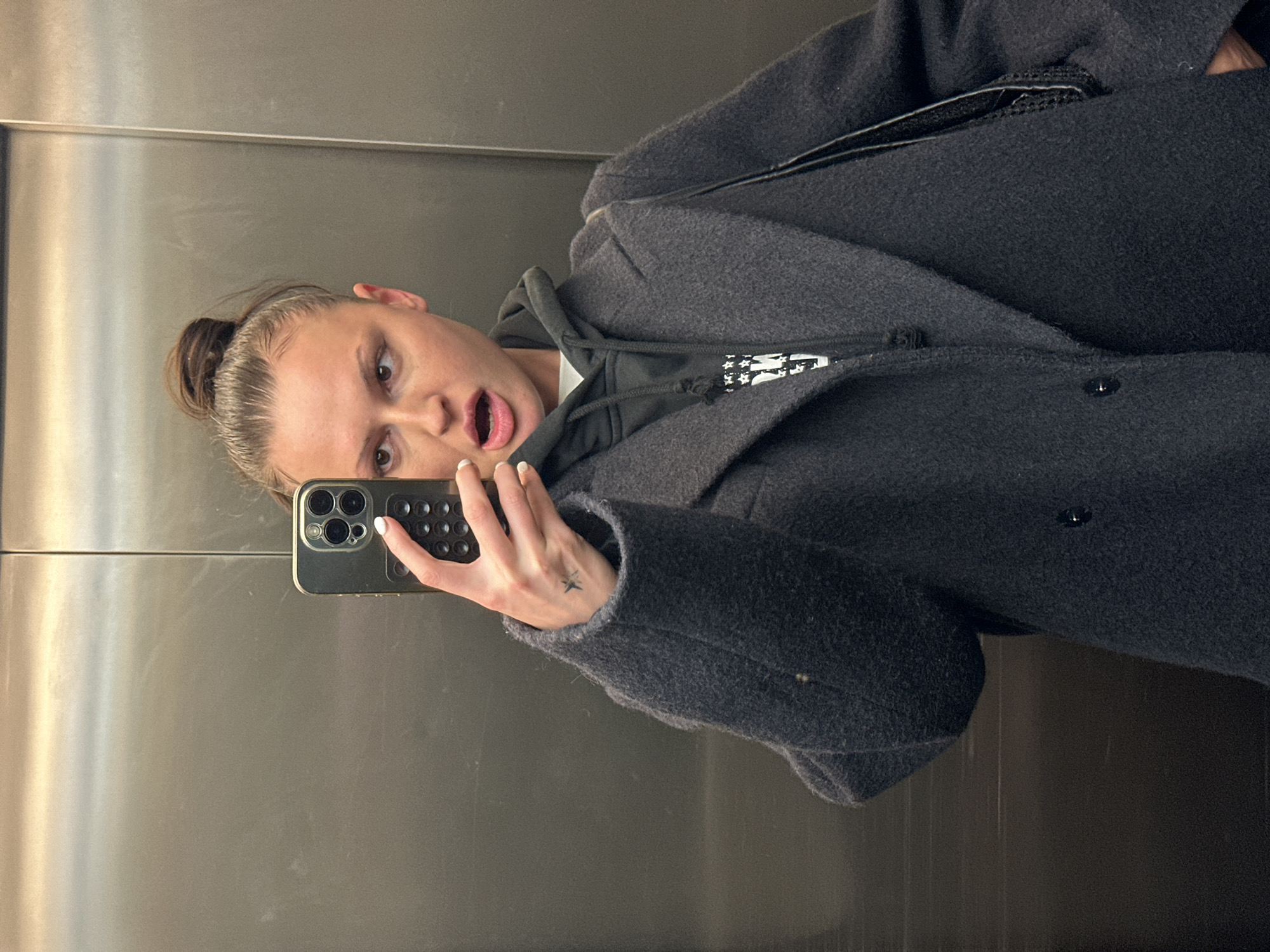 The image displays a female individual taking a selfie inside an elevator. The elevator interior features polished, metallic walls, likely stainless steel, with a vertical seam visible centrally behind the subject. The lighting is consistent with interior artificial illumination.
The subject is a light-skinned female with brown hair pulled into a high, messy bun. Her brown eyes are open, and her mouth is open in a slightly agape or surprised expression, revealing white teeth. She is dressed in a dark charcoal or navy blue textured overcoat, worn unbuttoned, over a dark hooded sweatshirt. A portion of a white garment is visible beneath the hoodie. A black strap, consistent with a handbag or shoulder bag, is visible over her left shoulder (viewer's right). Her right hand, adorned with white-painted fingernails, holds a dark-colored smartphone with multiple rear camera lenses, angled to capture the image. A small, dark, star-shaped tattoo is present on the back of her right hand, near the base of the index finger.
The specific location within the city of London, United Kingdom, cannot be determined from the visual information provided in the image.