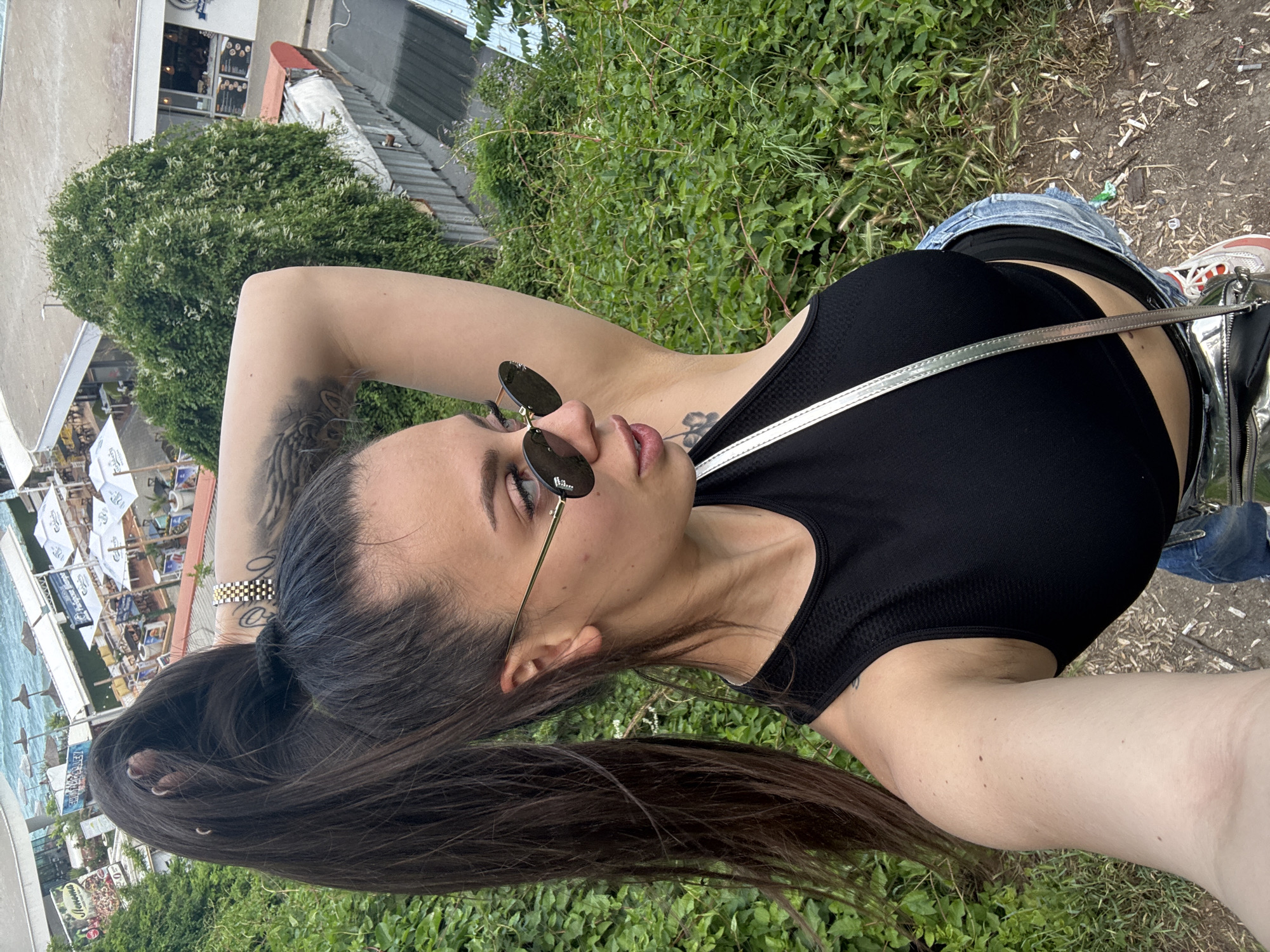 The image captures a young woman posing outdoors in a bright, daytime setting, likely in Varna, Bulgaria. She has long, dark hair tied in a ponytail and is looking upwards and to the right, wearing black sunglasses and a confident expression. Her attire includes a black athletic crop top and blue denim shorts or jeans. She has visible tattoos, including a winged design on her left upper arm and a floral motif near her chest. A gold and black checkerboard bracelet adorns her left wrist, and a silver strap from a crossbody bag crosses her chest.
The background reveals a vibrant coastal scene. To the left (when the image is correctly oriented), numerous white beach umbrellas, sunbeds, and the blue sea are visible, suggesting a lively beach resort area. Buildings, possibly cafes or shops, line the beach, with one sign clearly displaying "MENU" and the Bulgarian word "Vkusno" (delicious). The immediate surroundings are filled with dense green foliage and ground cover, indicating a natural, vegetated environment. The ground underfoot is a mix of dirt and sparse grass with some natural debris. The clear lighting indicates it is daytime, and the overall mood is casual and summery.