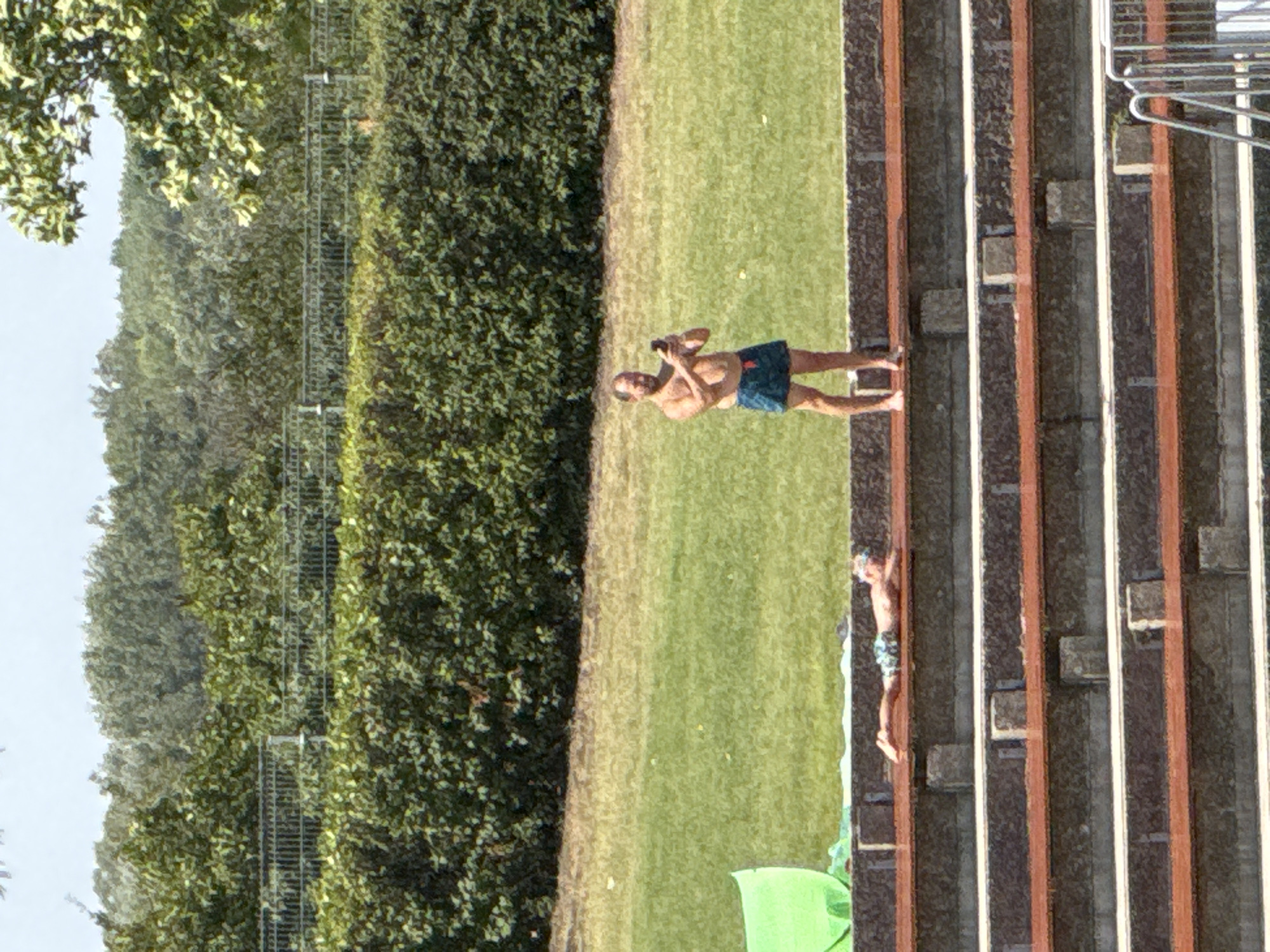 In Hainburg an der Donau, Austria, on a sunny day, two individuals are present in an outdoor recreational area. In the foreground, a person lies prone on a tiered seating structure, appearing to sunbathe. They are wearing patterned swim trunks and goggles. Further back and to the right, another individual stands upright, shirtless, and is holding a mobile phone, seemingly taking a picture or video. This person is wearing dark blue swim trunks. The environment features lush green grass, dense green foliage forming a hedge in the background, and a clear sky. The tiered seating is made of concrete and wood. A metal railing and stairs are partially visible in the lower right corner, suggesting the presence of a pool or similar water feature.