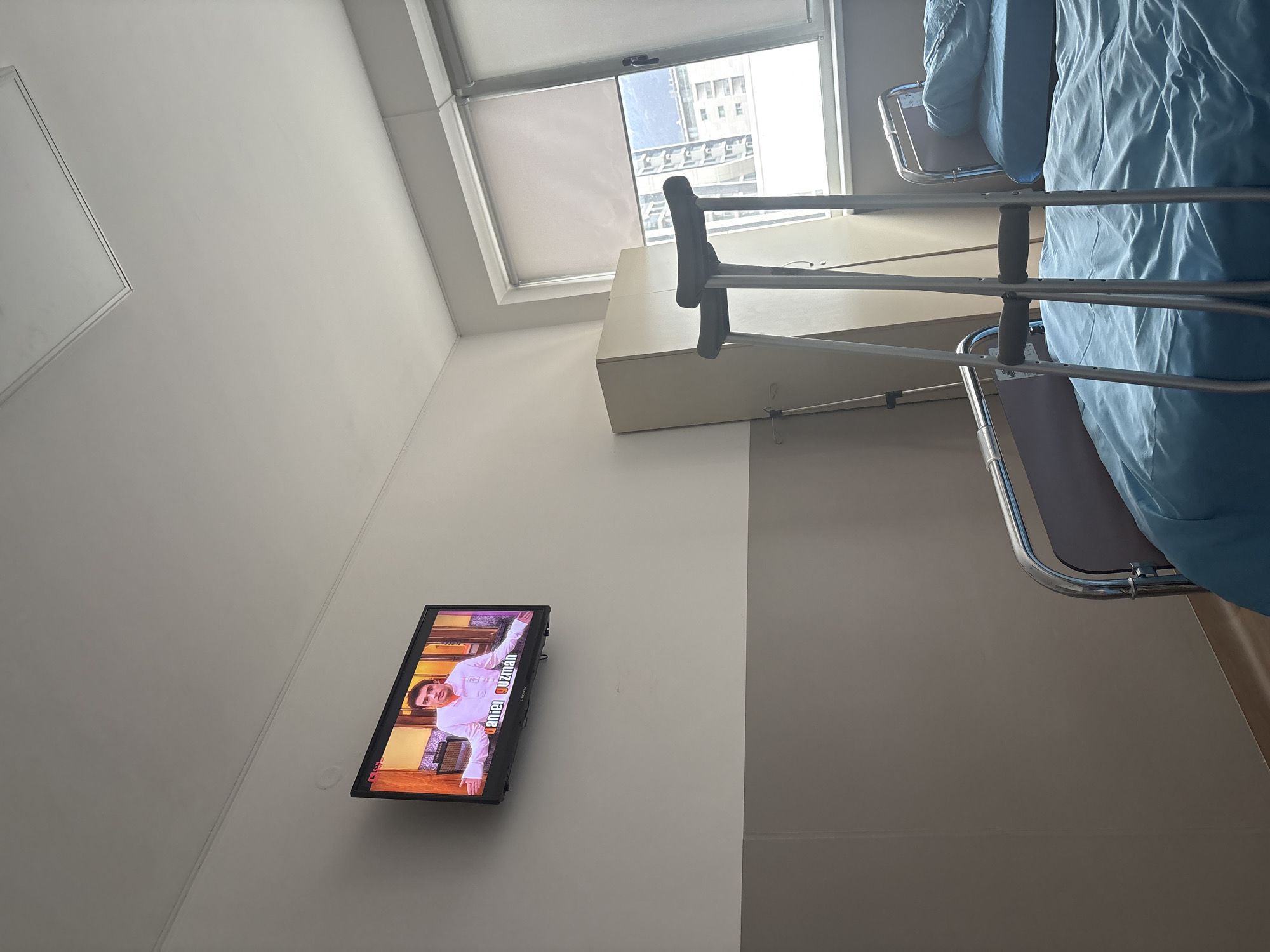 A hospital room in Sofia, Bulgaria, contains a bed draped with light blue sheets, a pair of crutches leaning against a bedside cabinet, and a wall-mounted television displaying a person named "Daniel Guzhan" on the screen. Outside the window are buildings that appear to be urban residential or commercial structures.