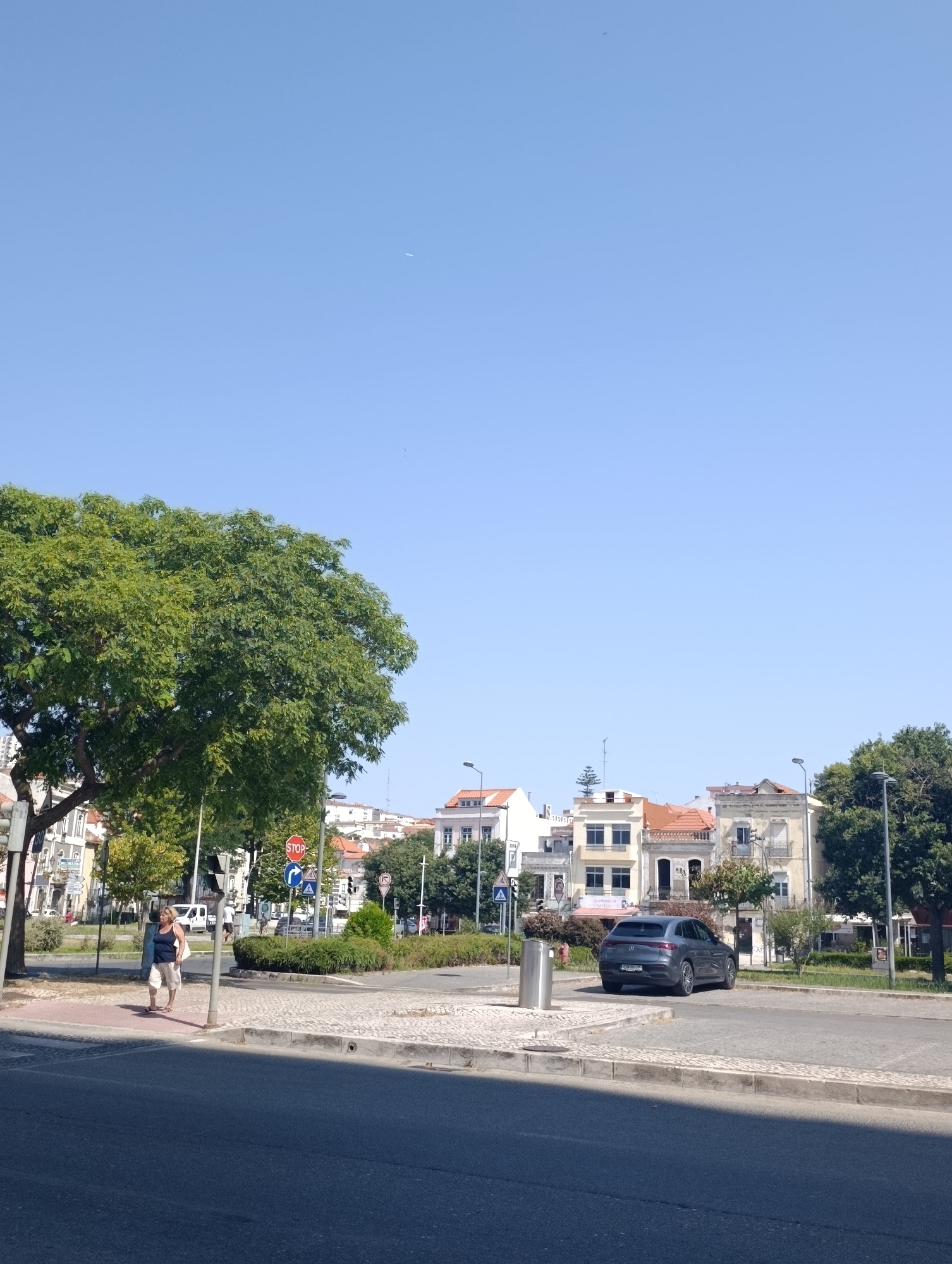 An eye-level, medium shot captures a sunlit street scene in Setúbal, Portugal, on a clear blue day. A large, leafy green tree dominates the left side of the frame, casting a shadow. In the foreground, a woman in a dark blue tank top and light-colored shorts walks with a white bag over her shoulder. She is on a sidewalk adjacent to a paved road. A gray car is parked to the right of the scene, facing away from the viewer. Behind the parked car, several buildings with light-colored facades and terracotta roofs are visible. Traffic signs, including a red stop sign and blue directional signs, are present at an intersection. The environment appears to be a moderately busy urban area.