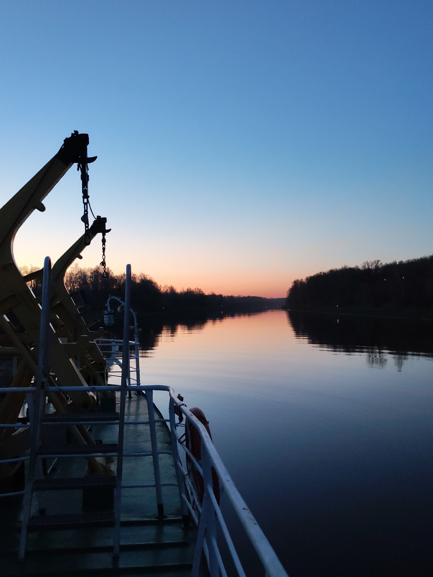 A dusk landscape captured from a boat on the canal named after Moscow, Russia. The sky is blue, fading to a vivid orange at the horizon. Silhouetted trees line both sides of the waterway. The water reflects the colors of the sky and the outline of the opposite bank. The boat's railing is visible on the left, as are parts of a crane structure. No people are visible.