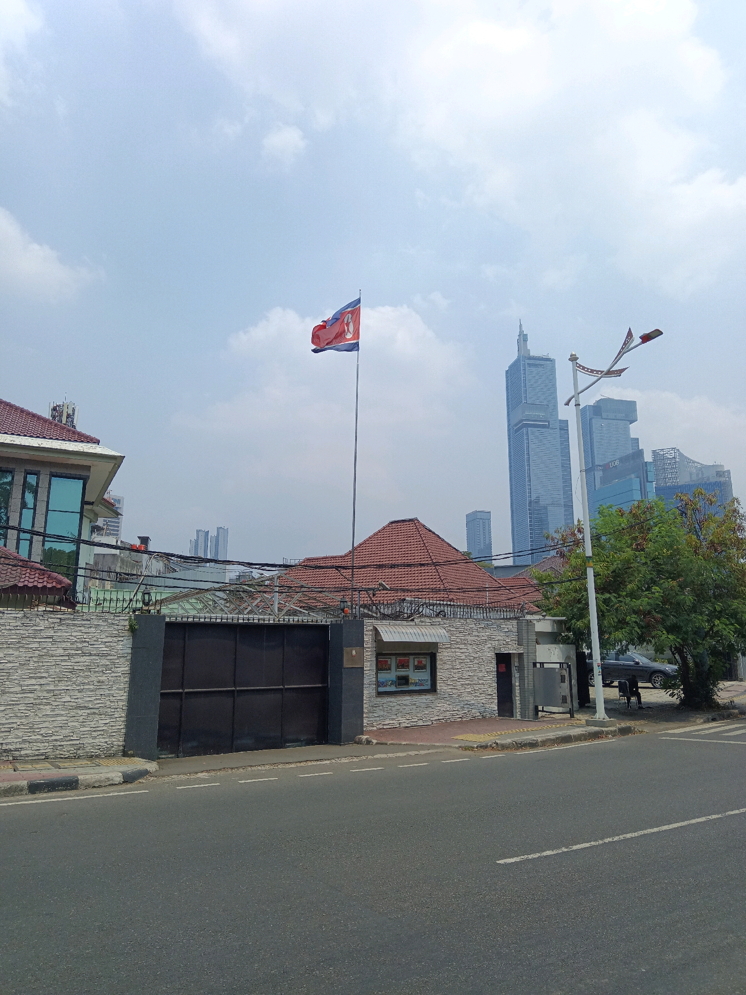 The image depicts the Embassy of North Korea in South Jakarta, Indonesia, under a partly cloudy sky. A large, dark metal gate, flanked by a stone wall, dominates the foreground on the left. A flagpole stands near the center, displaying the flag of North Korea. Behind the wall, a building with a red-tiled roof and a modern extension with large glass windows is visible. A small building with a window displaying pamphlets is situated to the right of the gate, adjacent to a street lamp adorned with small decorative flags. Further in the background, a dense cluster of modern skyscrapers pierces the sky, signifying the urban environment of Jakarta. A dark-colored car is parked near a large tree on the right side of the street. The street in the foreground is paved and shows white lane markings. There are no people clearly visible in the scene, though a figure can be discerned sitting on a chair near the parked car. The overall scene is static and captures a moment in time at this diplomatic location.