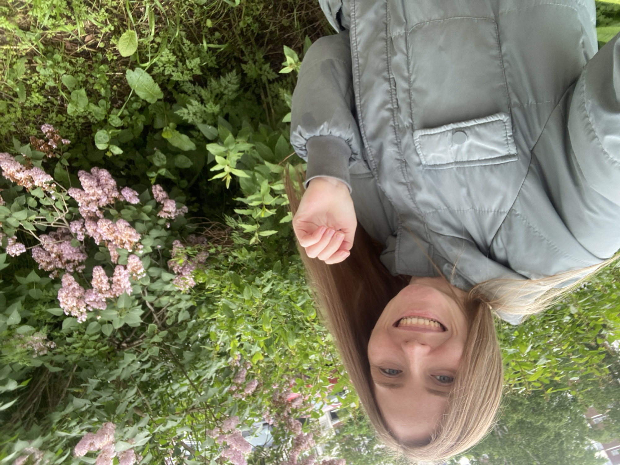 A young woman with long, blonde hair is taking a selfie in front of a blooming lilac bush. She is wearing a light grey, quilted jacket with a chest pocket. Her left hand is raised and her fingers are slightly curled. The background shows lush green foliage and clusters of pinkish-purple lilac flowers. Behind the bushes, parts of a building and a red vehicle are visible. The scene appears to be outdoors in a residential area, possibly in Yuzhno-Sakhalinsk, Russia, given the presence of lilac bushes, common in the region during bloom season.