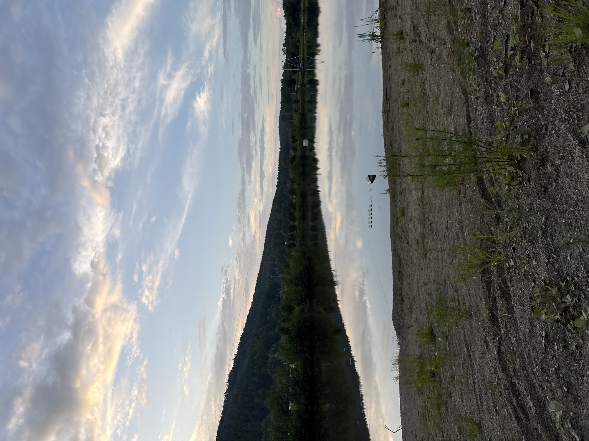 The image depicts a serene lakefront scene at dusk in Syrostan, Russia. The sky is filled with a mix of soft clouds, tinged with hints of yellow and orange from the setting sun, against a pale blue expanse. A dense, tree-covered hillside forms the backdrop on the left side of the image, with a few structures visible amongst the trees. The calm lake mirrors the sky and the distant landscape with remarkable clarity. In the foreground, a sandy, rocky shore is sparsely populated with green vegetation and tire tracks imprinted in the soil. Several floating structures, possibly for water sports or recreation, are visible on the surface of the lake, along with a distant tower-like structure with cables extending from it. The overall atmosphere is peaceful and natural.