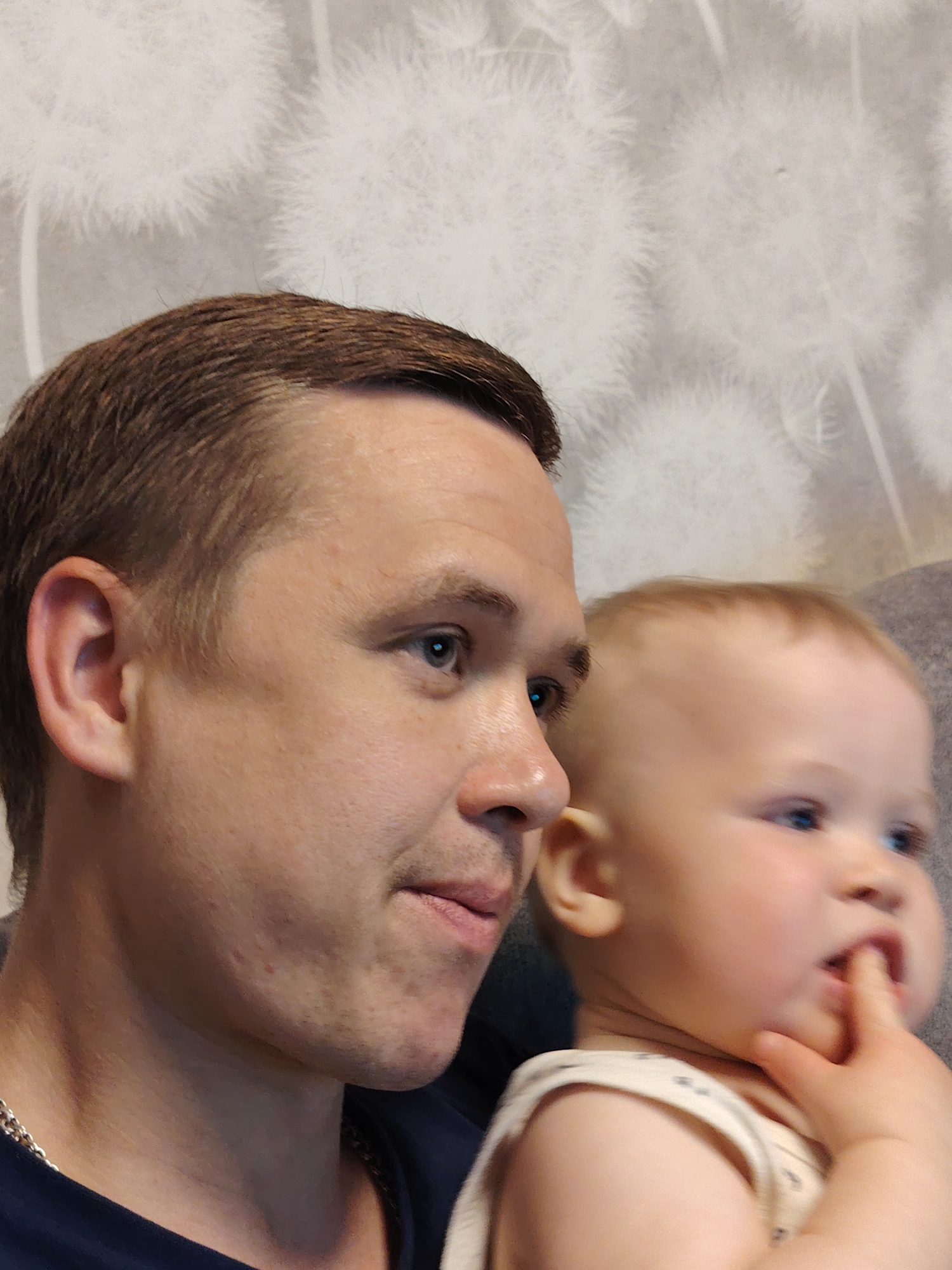 An indoor, eye-level, close-up photograph features a man and a baby, likely a father and child, in Chaikovsky, Russia. The man, with short brown hair, fair skin, and blue eyes, is in the foreground on the left, looking off-camera to the right. He has some stubble on his jawline and a few minor blemishes on his face and neck. He wears a dark blue t-shirt and a silver chain around his neck.
Behind and to the right of the man is a baby with light hair and blue eyes, also looking in the same direction. The baby has fair skin and chubby cheeks, with one hand bringing a finger to its mouth. Both subjects appear calm and engaged with something out of frame.
The background is a white wall with a subtle, textured pattern resembling fluffy dandelions or similar seed pods, suggesting wallpaper. The lighting is soft and even, typical of indoor conditions, highlighting the subjects without harsh shadows. No text is visible in the image.