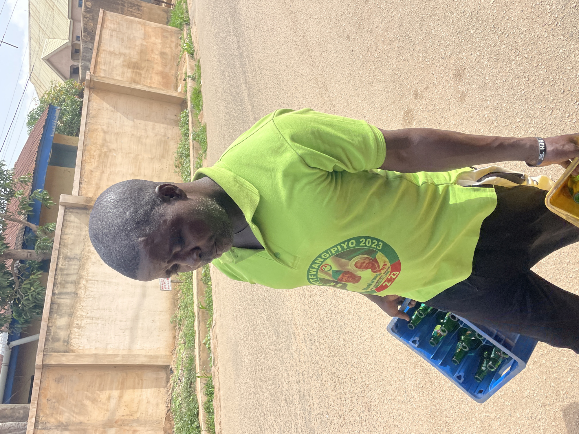 A man is walking along a street. He is carrying a blue plastic crate filled with green beer bottles. He is wearing a light green polo shirt with the words "Mutfwang/Piyo 2023" and an image of two people printed on it. He is also wearing black pants and a black wristband on his right wrist. The street is paved and there is a light-colored wall along one side of the street with green vegetation growing at its base. Buildings can be seen in the background. The scene is likely taking place in Jos, Nigeria, based on the text on the shirt.