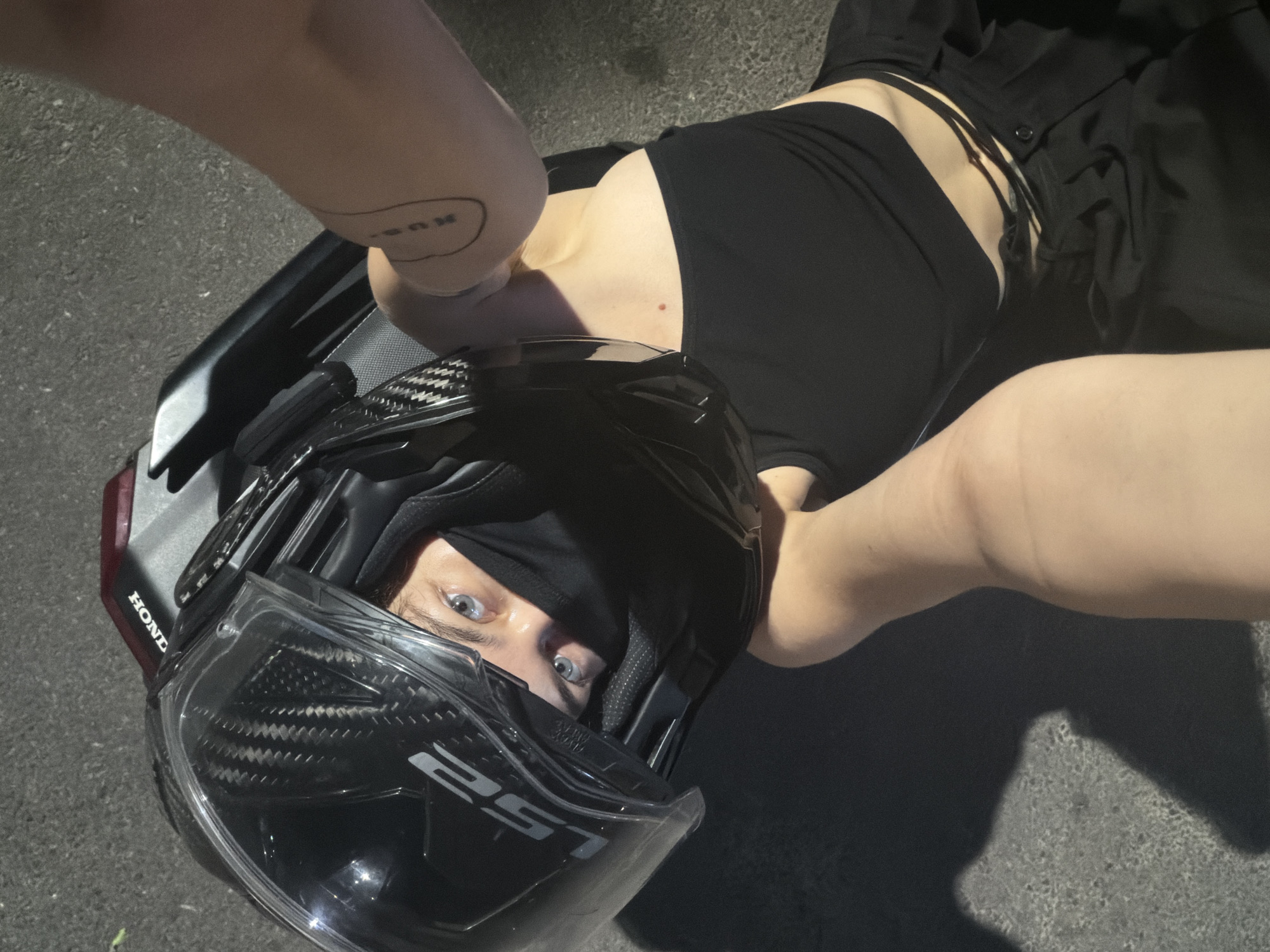 An overhead photograph captures a person lying on their back on a dark asphalt or concrete surface, likely outdoors in Đà Nẵng, Vietnam, during the evening or night. The individual wears a black sleeveless top with strap details on the side, and dark trousers or shorts.
Their head is encased in a black full-face LS2 motorcycle helmet, which features a visible carbon fiber pattern. A black balaclava or neck gaiter covers their mouth and nose, revealing only their direct, bright blue eyes, which gaze intently at the viewer.
Visible on the upper left arm is a tattoo, appearing as stylized text within an irregular oval shape. To the upper left of the helmet, a red/maroon section of what appears to be a motorcycle or scooter fairing is visible, bearing the word "HONDA." The scene is dimly lit, with artificial light highlighting the person's torso and parts of the helmet, while the background remains dark. The overall impression is a casual, perhaps edgy, portrait, possibly taken during a break from motorcycling.