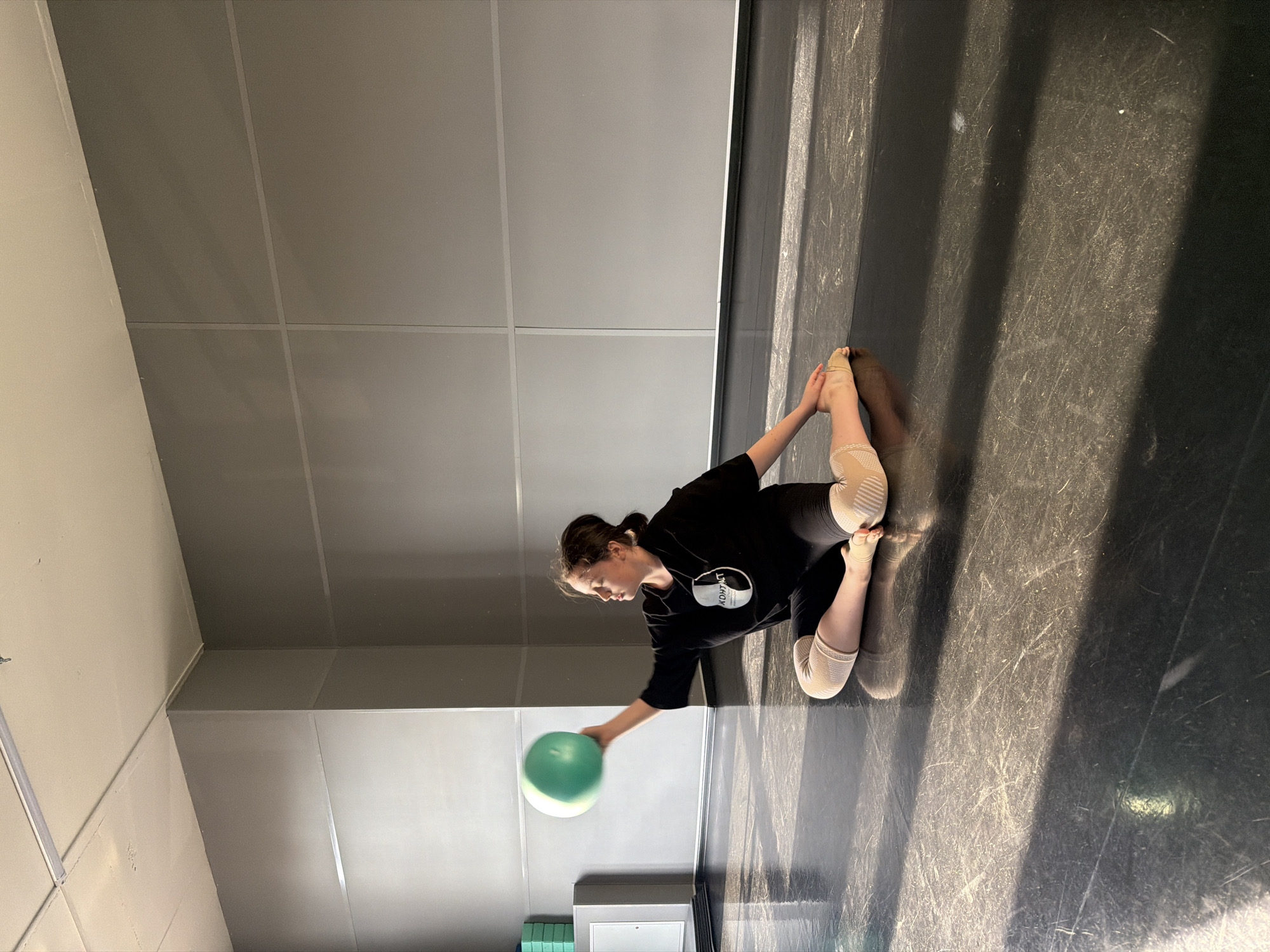 A young female gymnast is shown sitting on a dark floor in a room with light-colored walls. She is wearing a black long-sleeved shirt, black leggings, and leg warmers. Her hair is pulled back in a ponytail. She is holding a light green ball in her right hand. The floor appears to be a performance space. The room is well-lit with sunlight streaming in from the right. There is equipment in the bottom center of the frame. The location is likely a gymnasium or dance studio in Saint Petersburg, Russia, based on the user's request.