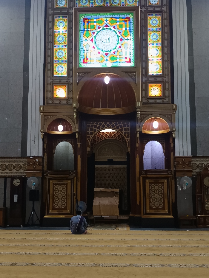 An interior view of Masjid Agung Cianjur (Great Mosque of Cianjur) in Cianjur, Indonesia, shows a single individual seated on a patterned light beige carpet, facing an ornate wooden and stained-glass prayer wall. The person is observed from the rear, wearing a light blue-grey shirt and dark trousers, with a purple-striped sash draped over their left shoulder and back, sitting in a posture indicative of prayer or contemplation.
The dominant feature is the elaborately decorated wall at the front of the prayer hall. It comprises a large central rectangular stained-glass window featuring complex geometric patterns in green, blue, yellow, red, and white, with the Arabic word "Allah" (الله) centrally inscribed. This is flanked by narrower vertical and smaller square stained-glass panels with floral and geometric motifs. Below these, the wall is constructed from dark wood with intricate carvings and golden accents. Two prominent wooden domes with integrated light fixtures illuminate the upper section. Further down, multiple arched alcoves and recessed areas are visible, some also illuminated. The central mihrab niche, appearing dark, is positioned at the base of the ornate structure, with a brown, draped object, possibly a lectern or covered furnishing, positioned in front of it.
On the left side of the scene, near the wall, a blue pedestal fan and a black speaker on a tripod stand are visible, alongside a wall-mounted clock. On the right, another blue pedestal fan and a similar wall clock are present. The room is dimly lit, with focal points of light emanating from within the ornate wall structure and the stained-glass panels. The floor is covered by a light-colored carpet featuring darker patterned stripes extending towards the foreground.