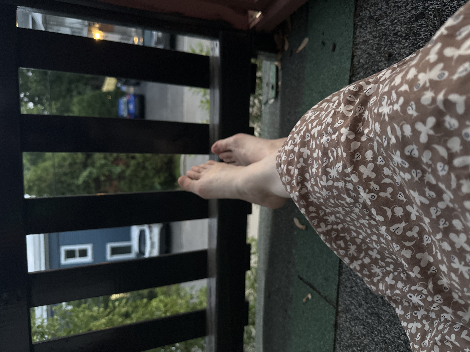 The image shows a first-person perspective from a balcony or porch in McMasterville, Canada. The foreground is dominated by the lower half of a person wearing a brown dress with a white floral pattern, and their bare feet are resting on a dark railing. Beyond the railing, a street scene is visible. A blue house with white-trimmed windows is on the left, and a white car is parked in front of it. Further down the street, a blue car is driving away. Trees and other houses are visible in the background. The lighting suggests it is either dusk or an overcast day.