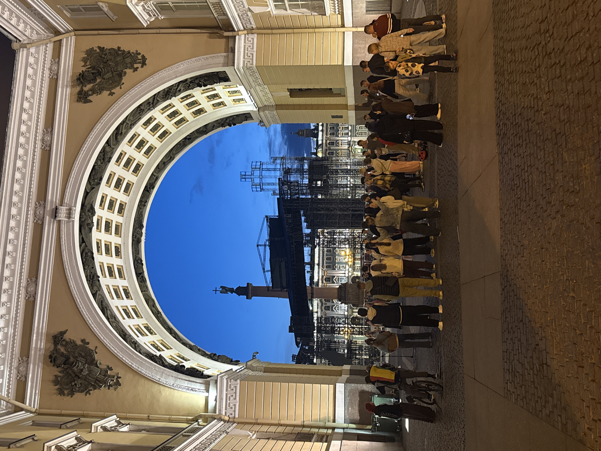 The image depicts a nighttime scene under a large archway, likely in Saint Petersburg, Russia, given the architectural style and the iconic Alexander Column visible in the background. A crowd of people is gathered in the open area beneath and beyond the arch.
The archway itself is grand, with ornate carvings and decorative elements. It frames a view of a large, open square, paved with cobblestones. In the distance, the Alexander Column, topped with an angel, stands tall, a prominent landmark. Surrounding the column are buildings and what appears to be a stage or construction scaffolding, suggesting an event or ongoing development.
The people in the foreground and midground are diverse in age and attire. They are standing, some in groups, others individually, and appear to be observing the scene or interacting with each other. One person on the left is near a bicycle, and another person further back is holding a flag. Some individuals are holding up their phones, possibly taking pictures or videos. The lighting suggests it is dusk or early night, with artificial lights illuminating the architecture and the gathering. The sky is a deep blue, indicating the twilight hours.