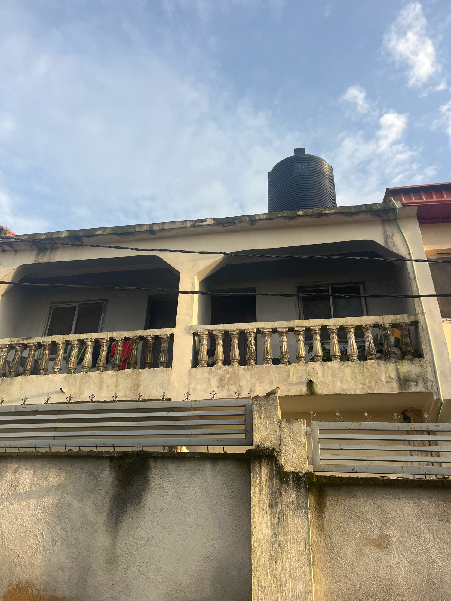 The image depicts a two-story building with a balcony on the upper level. The building is painted in a light color and shows signs of wear, with some discoloration and stains on the exterior walls. A large black water tank is situated on the flat roof of the building. The balcony is adorned with ornate railings, and some clothing items are hanging on them. Below the balcony, a fence separates the property from the street. A bird is perched on a pillar of the fence. The sky is partly cloudy, with a mix of blue sky and white clouds. The lighting suggests it is either early morning or late afternoon. The overall scene is typical of a residential area in Jos, Nigeria, characterized by its architectural style and the presence of essential utilities like water tanks.