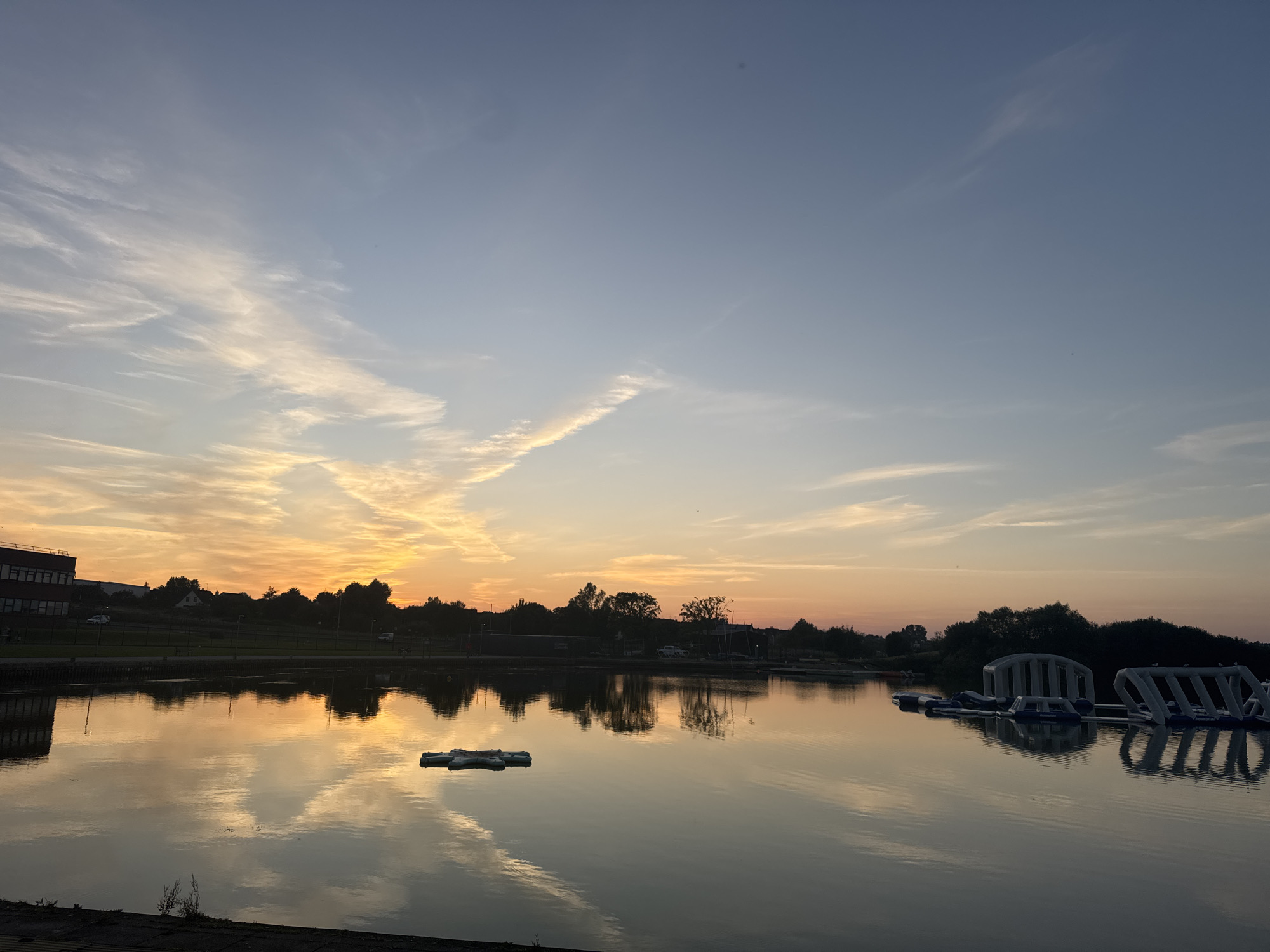 The image captures a serene sunset over the Craigavon Lakes in Craigavon, United Kingdom. The sky displays a gradient of soft blues and oranges, with wispy clouds illuminated by the setting sun. The water's surface reflects the vibrant colors of the sky and the silhouettes of trees and buildings lining the shore. In the foreground, a body of water stretches across the frame, with ripples indicating a gentle breeze. A few floating inflatable structures, commonly found in water parks, are visible on the right side of the lake. On the left, a multi-story building with numerous windows stands beside a tree-lined embankment. The overall atmosphere is tranquil and picturesque, showcasing a typical evening scene at this recreational area.