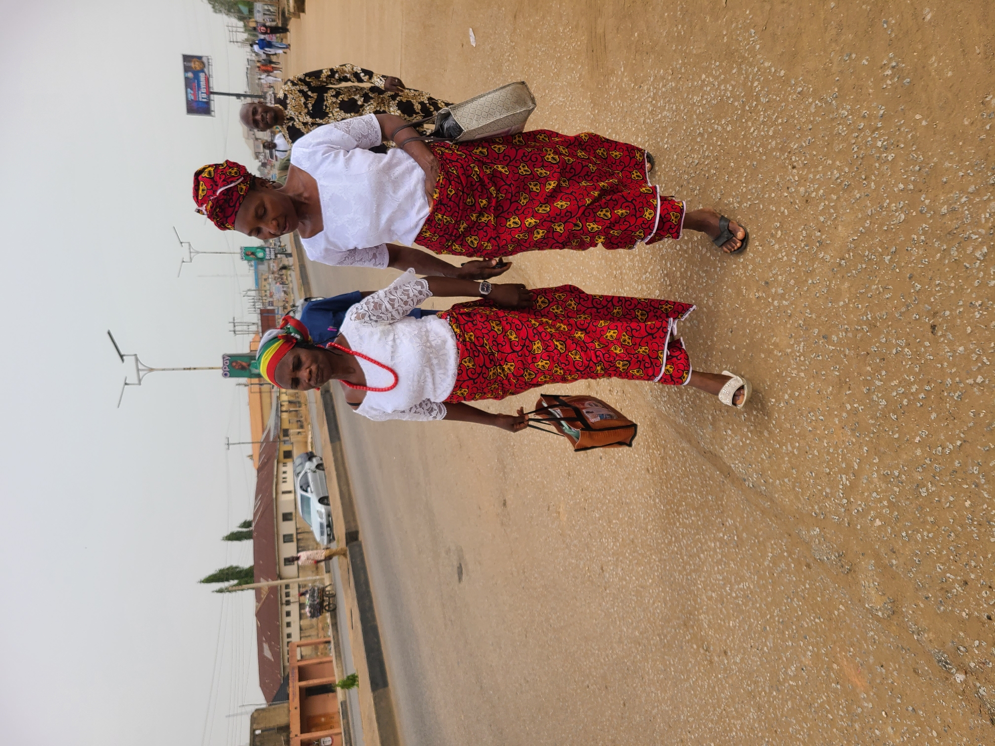 The image, taken in Jos, Nigeria, depicts an outdoor scene on what appears to be a street or roadside, featuring several people walking. In the foreground, two women walk side-by-side, moving towards the right of the frame. Both are dressed in white lace tops and striking red skirts patterned with yellow and black designs, a common African print. Each woman also wears a distinct head wrap: one in red with yellow patterns, and the other featuring stripes of red, yellow, and green.
The woman in the immediate foreground has a red beaded necklace and carries a small brown bag. Her companion carries a grey handbag slung over her shoulder and wears a watch. Both are wearing sandals or slippers. Further back, another woman in a black and gold patterned outfit is partially visible, along with a man whose head is seen. More people and vehicles are discernible in the distance along the road.
The ground is a light brown, somewhat rough-textured surface, suggesting a paved road or shoulder with visible aggregates. Buildings of various architectural styles line the street in the background, including one with a white car parked nearby. Utility poles and streetlights are also present. The sky is overcast or hazy, indicating a bright daytime setting with diffused natural light. A billboard in the background displays the word "OPAY". The scene suggests everyday activity in an urban or semi-urban environment.
