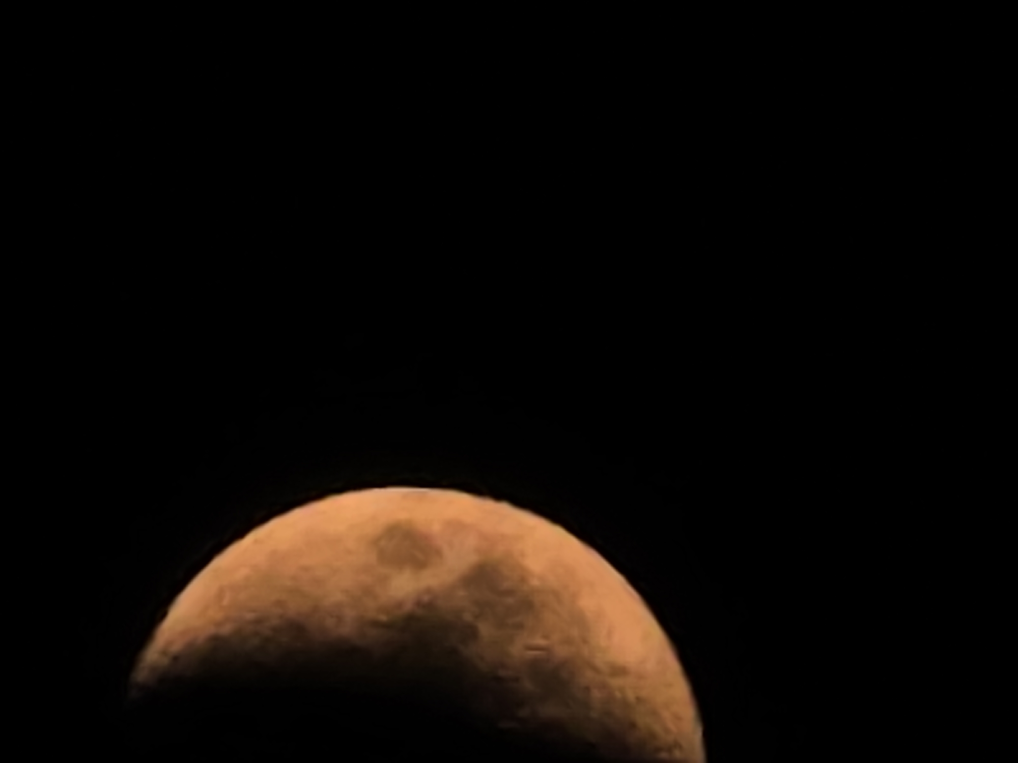 The image shows a close-up of the moon, with its crescent shape illuminated against a black background. The moon is a warm orange color, and its surface details, such as craters and darker maria, are visible. The image does not contain any information or context related to the city of Yambol, Bulgaria, or any urban environment.