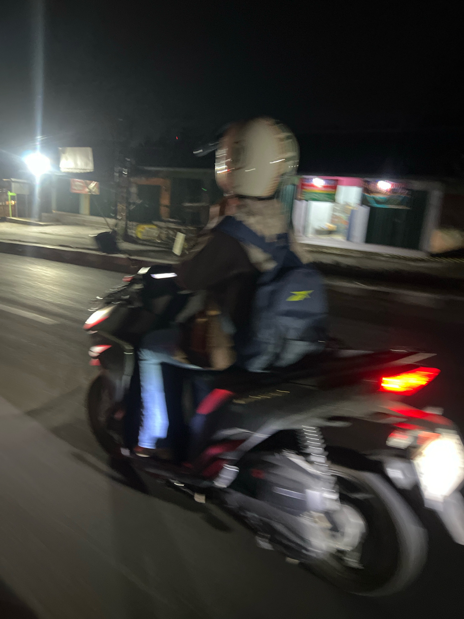The image depicts a person riding a motorcycle at night on a road, likely in Salam, Indonesia, given the presence of buildings and street lighting consistent with the region. The motorcycle's brake lights are illuminated, indicating it is either braking or in motion with the lights on. The rider is wearing a helmet and a backpack. The surroundings are dimly lit, with some buildings and structures visible in the background, suggesting an urban or semi-urban environment. The motion blur in the image indicates that the photograph was taken while the motorcycle was moving.