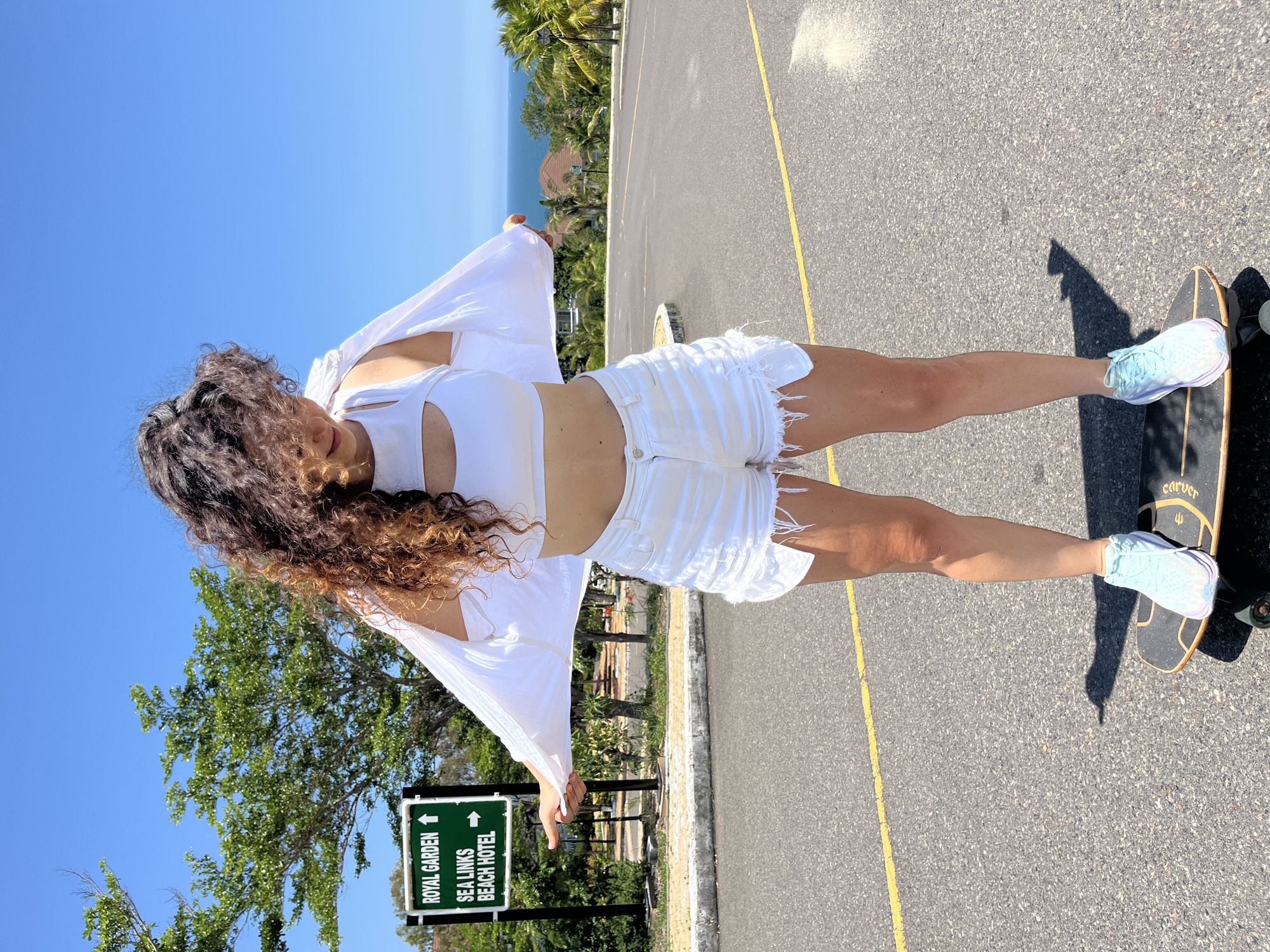 A young woman with long, curly brown hair is standing on a skateboard on an asphalt road under a clear blue sky. She is wearing a white crop top, white ripped denim shorts, and light blue sneakers. She is holding a white open-front cardigan with both hands, letting it drape over her arms. Behind her, a green directional sign indicates "ROYAL GARDEN" with an upward arrow and "SEA LINKS BEACH HOTEL" with a rightward arrow. Trees and palm trees are visible in the background, with a body of water and distant buildings in the far distance. The scene appears to be in a tropical or coastal location. The specific location is the city of Phan Thiet, Vietnam.