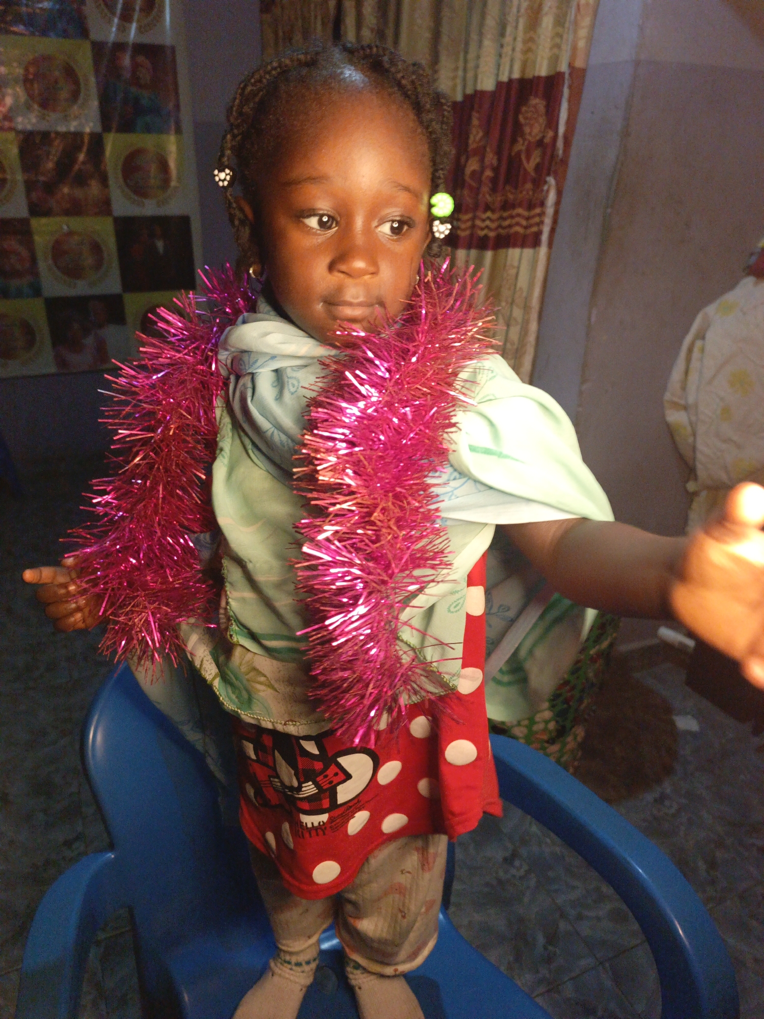 A young girl, likely of Nigerian descent, is positioned in the center of the frame, seated on a blue plastic chair. She has dark skin, braided hair, and is wearing a mint-green patterned shirt, a red and white polka dot skirt, and white socks. A bright pink tinsel garland is draped around her neck. Her left arm is extended slightly towards the viewer. The background features a tapestry or curtain with intricate patterns and a tiled wall. The overall lighting is warm, suggesting an indoor setting, possibly in Akwanga, Nigeria, given the context.