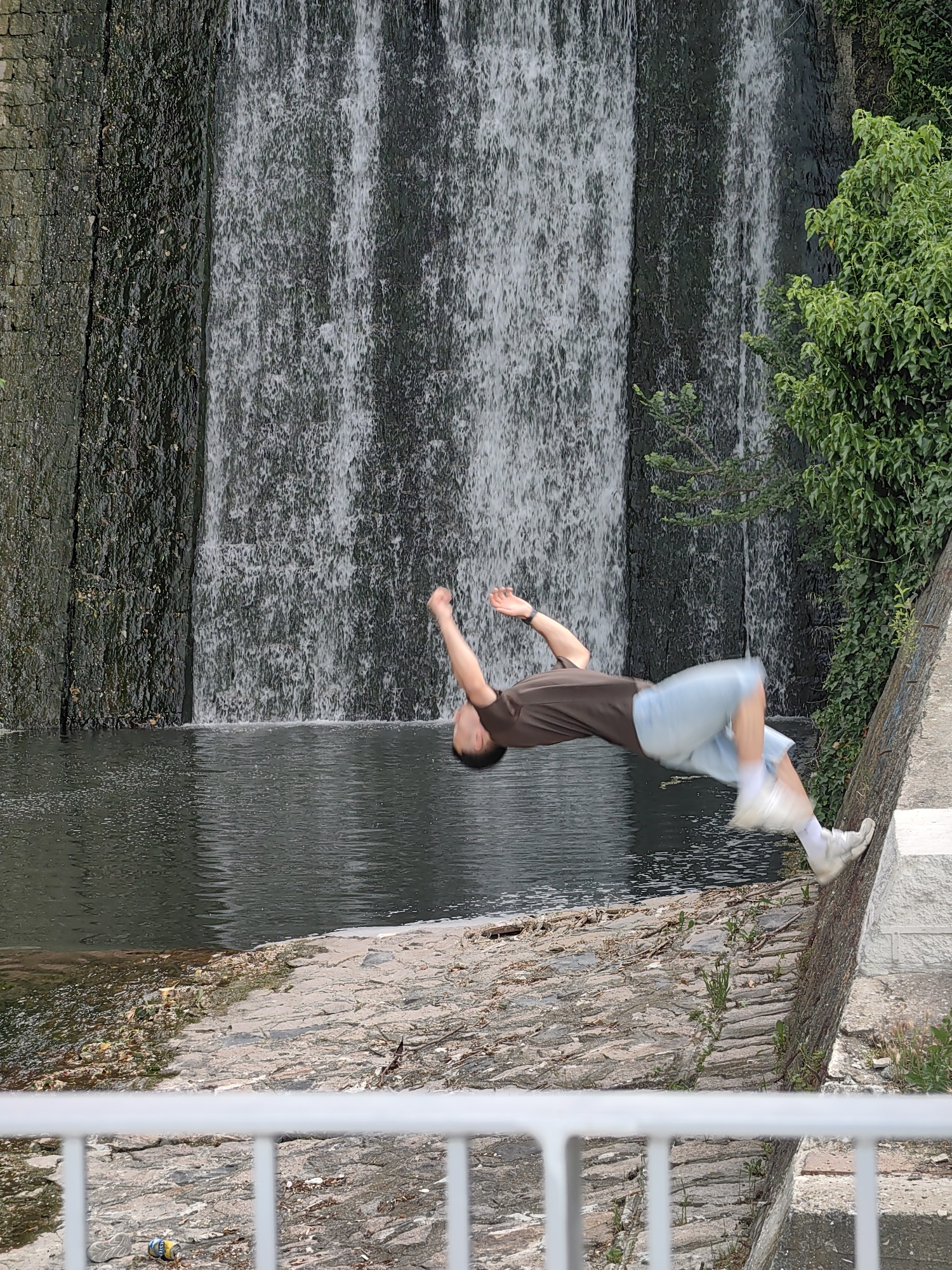 A man is performing a backflip near a waterfall in Varna, Bulgaria. He is wearing a brown t-shirt, light blue shorts, and white sneakers. The waterfall is made of stone and is surrounded by greenery. A white railing is in the foreground. Debris litters the ground near the railing.