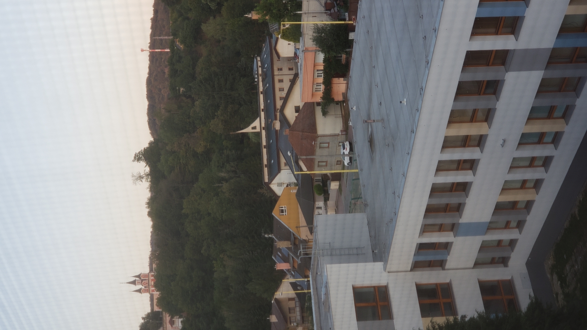 The image is an exterior, elevated view of a residential area and a school building in Prešov, Slovakia. The foreground features a modern, multi-story school building with a grey, flat roof and rows of identical windows with brown frames. The building's facade is primarily white, with accents of light blue and yellow panels between some window sections. To the left of the school, a portion of a larger, white building with large, square windows is visible.
In the middle ground, a street runs through a neighborhood of houses with pitched roofs of various colors, including yellow, orange, and brown. Some houses have visible chimneys, and streetlights with yellow poles are present. A white car is parked on the street. Beyond the houses, a dense forest covers a hillside.
In the background, atop the hill, a prominent red and white church with multiple spires can be seen. Further in the distance, a tall red and white telecommunications tower stands against the sky, which is a pale blue with a hazy quality, suggesting either early morning or late afternoon light. The overall impression is a quiet, suburban or semi-rural landscape within the city.