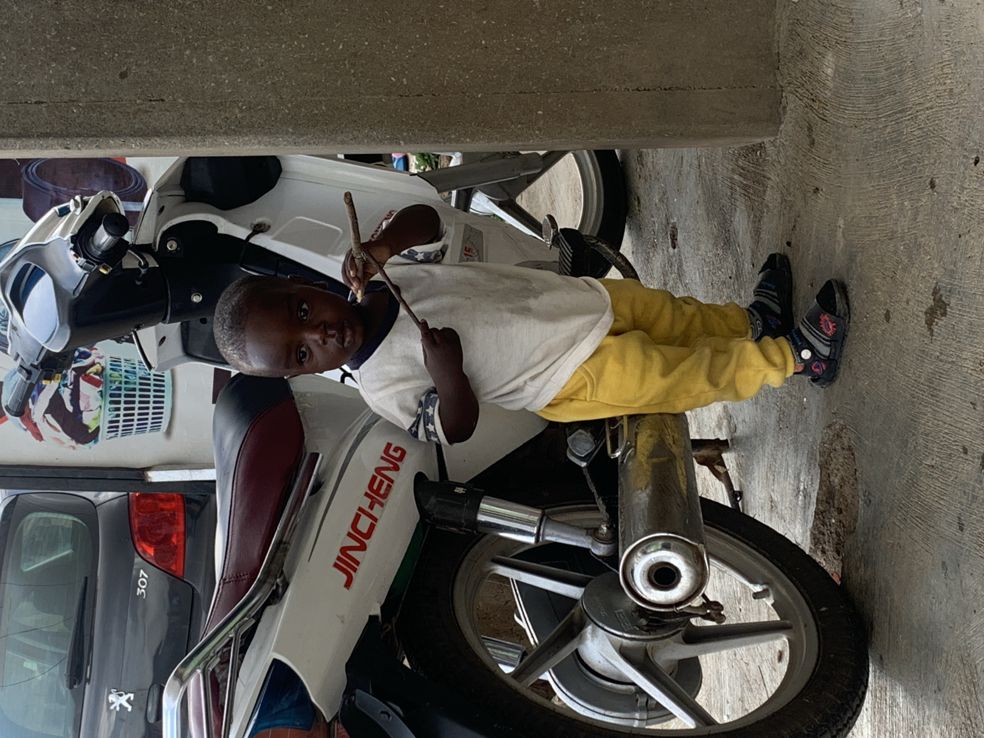In Jos, Nigeria, a young child stands next to a white motorcycle, holding a stick. The child is wearing a white t-shirt with blue trim on the sleeves and collar, and yellow pants. They are also wearing dark sandals with colorful accents. The motorcycle is partially visible, with a portion of the seat and the rear wheel showing. A dark-colored car, possibly a Peugeot 307, is parked behind the motorcycle. The ground is concrete, with some dirt and debris scattered. A concrete pillar is located to the right of the child. In the background, a white basket filled with colorful laundry is visible on a wall. The scene appears to be outdoors, possibly in a residential or commercial area.
