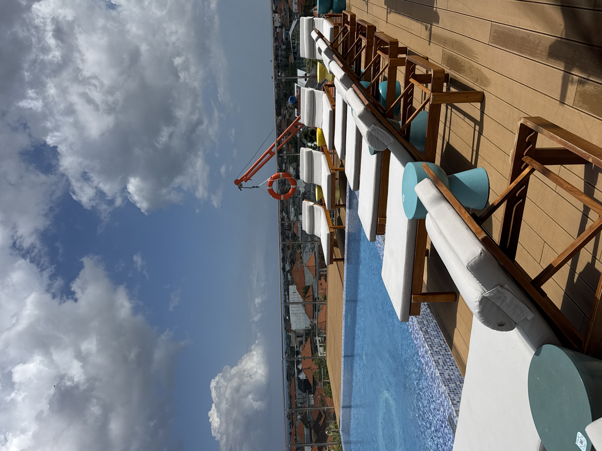 The image depicts a rooftop swimming pool area in North Kuta, Indonesia, bathed in daylight under a cloudy sky. A bright blue swimming pool with a mosaic tile border occupies the left side of the frame, reflecting the sky. Lined up on the wooden deck are several white lounge chairs, some with cushions, arranged in rows. Various small, colorful side tables, primarily teal and yellow, are interspersed between the lounge chairs. In the background, beyond a metal railing, a city skyline with terracotta rooftops and buildings is visible. A prominent orange construction crane with a life preserver attached stands in the middle ground. The deck itself is made of brown wooden planks, showing some wet marks and casting shadows from the lounge chairs.