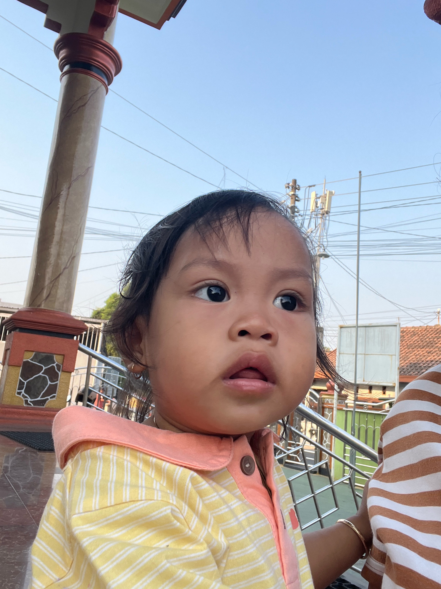 A young child with dark hair, dark eyes, and light skin is shown from the chest up, looking to the right of the frame. The child is wearing a yellow and white striped shirt with a pink collar and a button. The background shows a clear blue sky with power lines and a utility pole. To the left, there is a section of a decorative pillar with red and brown accents and a mosaic pattern. In the mid-ground, there is a metal railing, some trees, and buildings with orange-tiled roofs. The setting appears to be outdoors in a residential or public area in Kecamatan Pati, Indonesia.