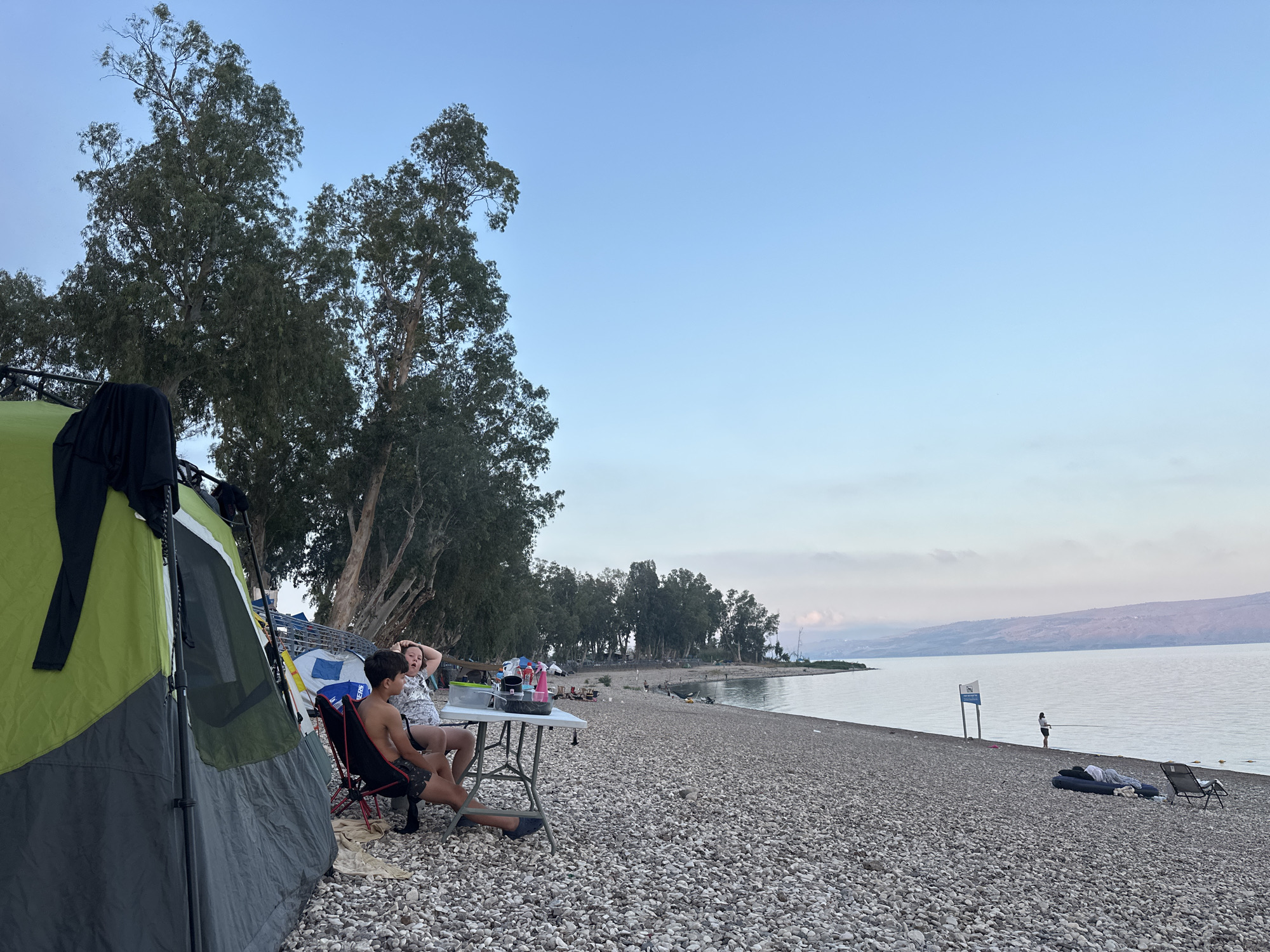 The image depicts a scene at a pebble beach on the shore of the Sea of Galilee in the Emek HaYarden Regional Council, Israel. A green and gray tent is pitched on the left side of the frame. Two children are seated at a white folding table to the right of the tent. The boy is wearing dark swimming shorts and is seated in a red camping chair, facing the camera. The girl, in a patterned top, is seated in a black camping chair, with her hands behind her head. A cooler and other items are on the table. Beyond the children, the pebble beach extends towards the calm water of the Sea of Galilee. A line of trees with thin trunks and sparse foliage runs along the edge of the beach, separating it from a more distant landscape. In the background, a person is standing on the shore, fishing, with some inflatables and chairs on the beach. The sky is clear and pale blue, with a hint of haze near the horizon, suggesting it is either early morning or late afternoon. The overall atmosphere is relaxed and suggests a camping or recreational outing.