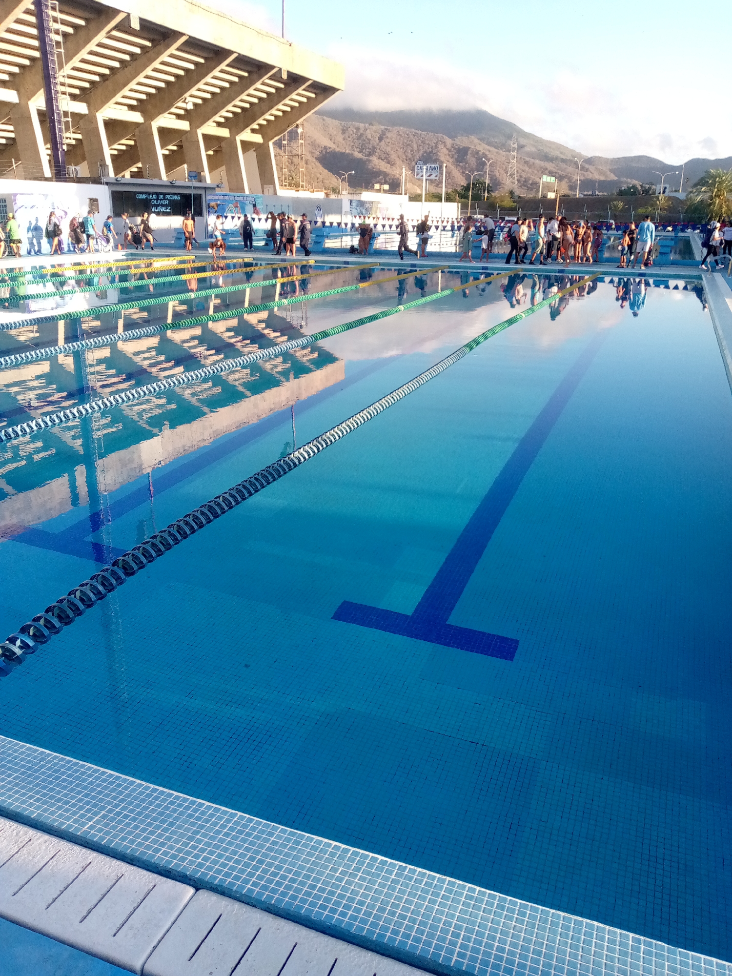 The image shows an outdoor swimming pool located in Caracas, Venezuela. The pool is rectangular and filled with blue water, with lane markers visible. A group of people is standing along the poolside. In the background, there is a stadium and mountains.